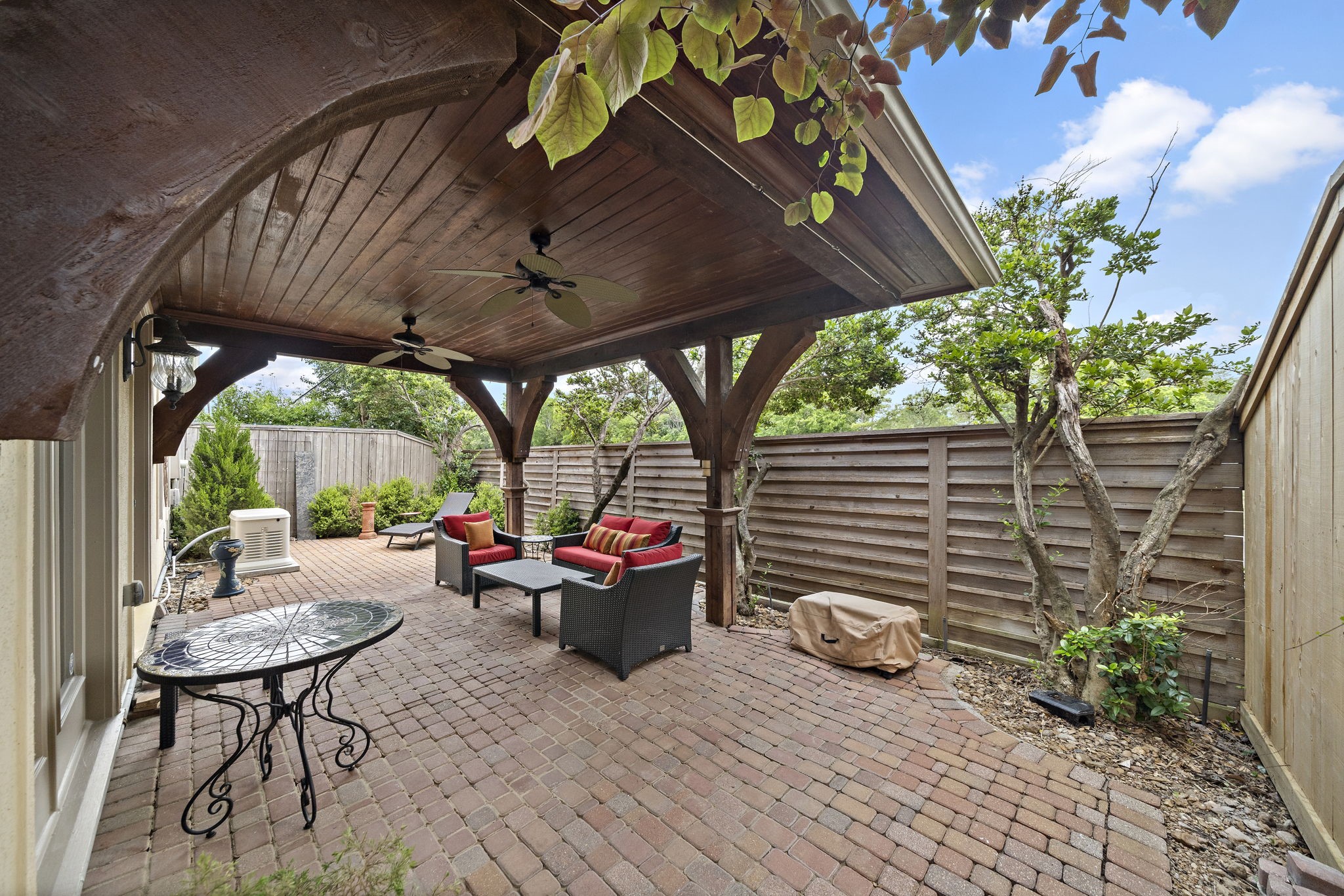 1229 Ripple Creek Drive Houston, TX 77057 - Photo 32 of 49 This photo showcases a charming outdoor patio with a brick floor and wooden ceiling. It features comfortable wicker seating with red cushions, ceiling fans, and decorative lighting, creating a cozy and inviting space for relaxation and entertaining.