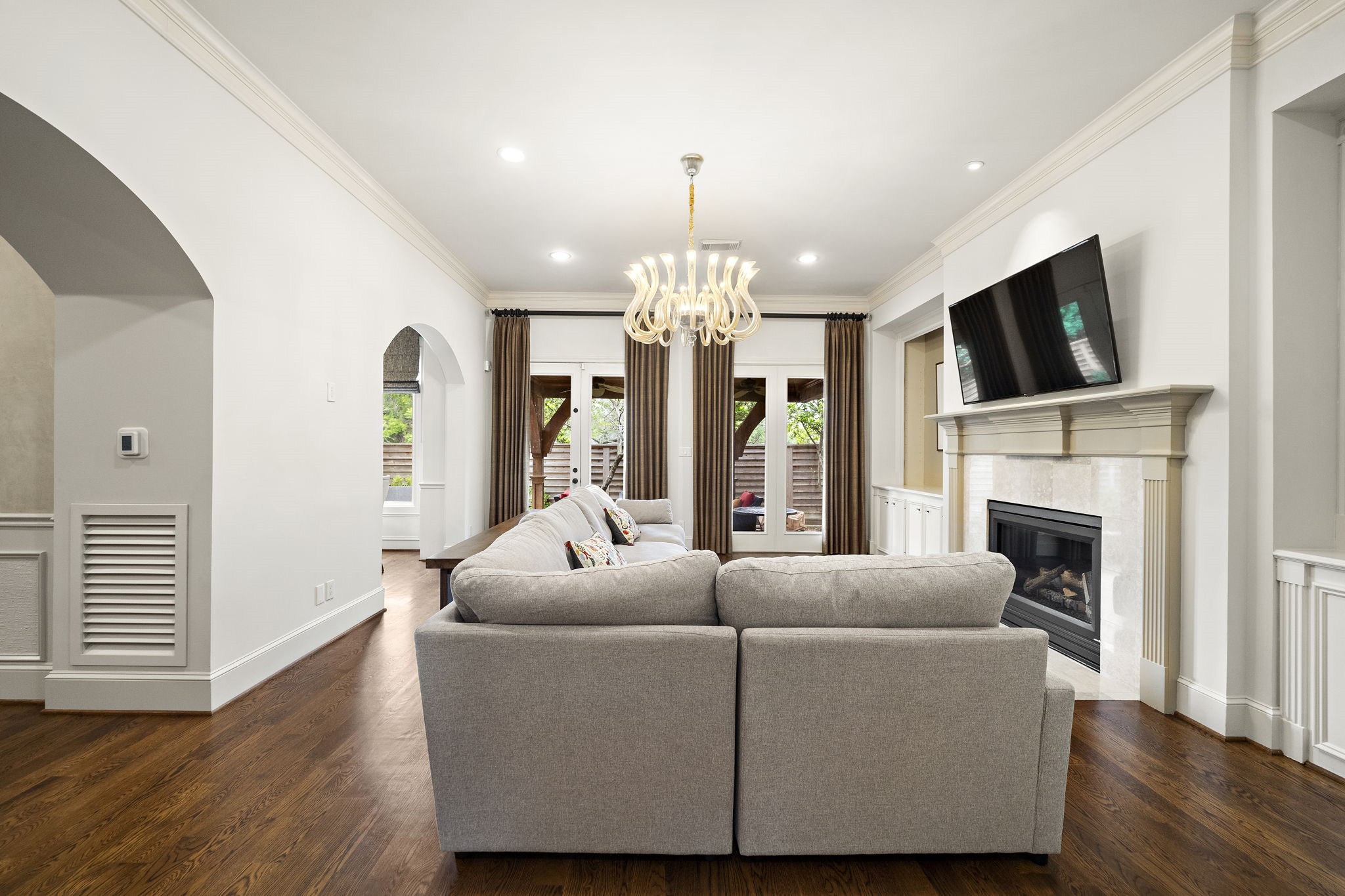 1229 Ripple Creek Drive Houston, TX 77057 - Photo 10 of 49 This living room features a cozy fireplace, mounted TV, and elegant chandelier. It has hardwood floors, a large gray sofa, and ample natural light from the windows and glass doors leading outside. The space feels open and inviting, perfect for relaxation or entertaining.