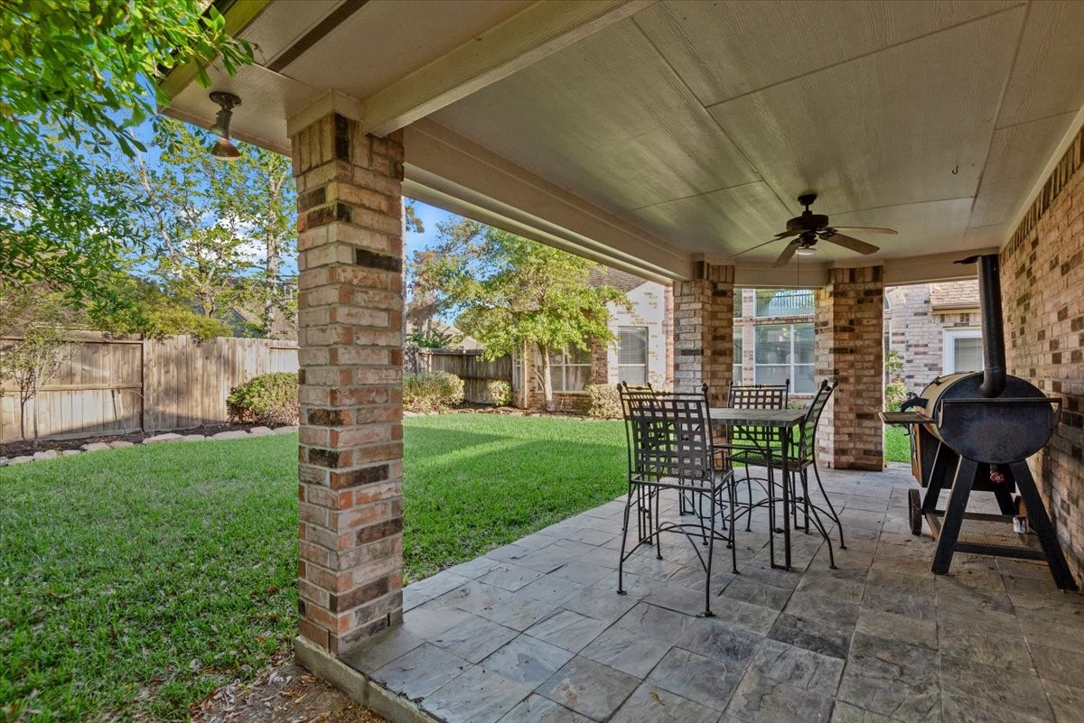 6507 Quiet Pointe Drive Spring, TX 77389 - Photo 29 of 46 This photo showcases a covered patio with brick pillars, featuring a ceiling fan and outdoor seating. It overlooks a spacious, well-maintained lawn with surrounding trees and a wooden fence, ideal for outdoor relaxation and entertainment.