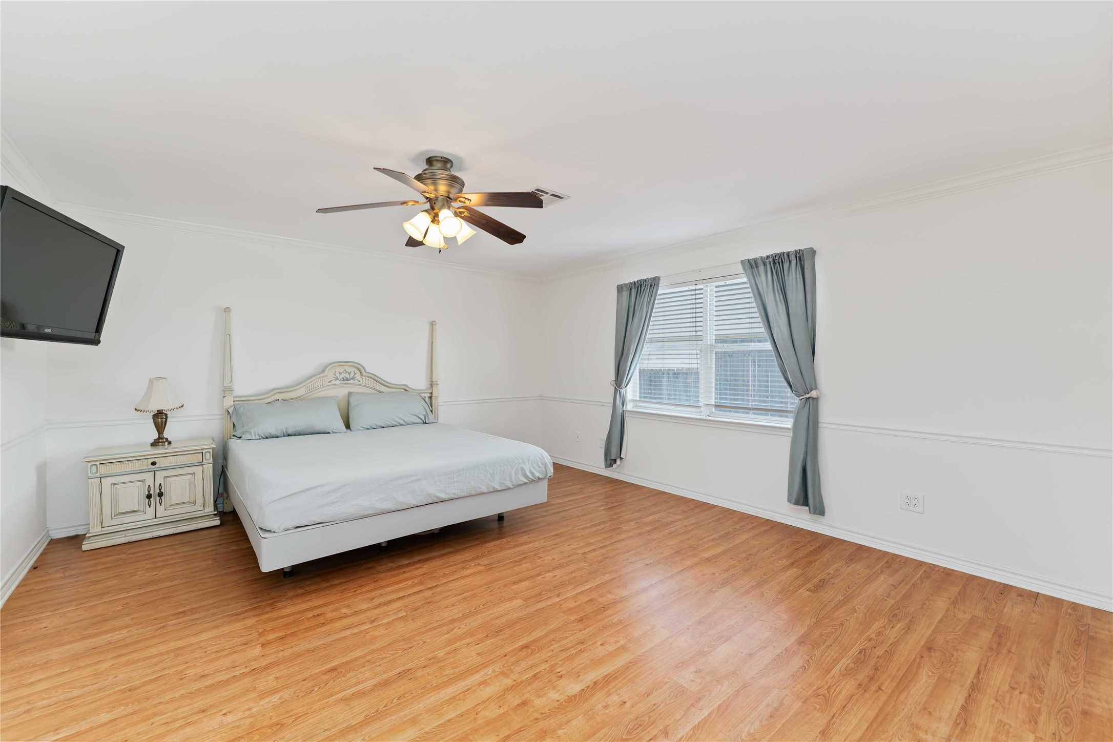 606 Packer Court Webster, TX 77598 - Photo 11 of 28 This primary bedroom features hardwood floors, a ceiling fan, and ample natural light from a large window dressed with curtains.