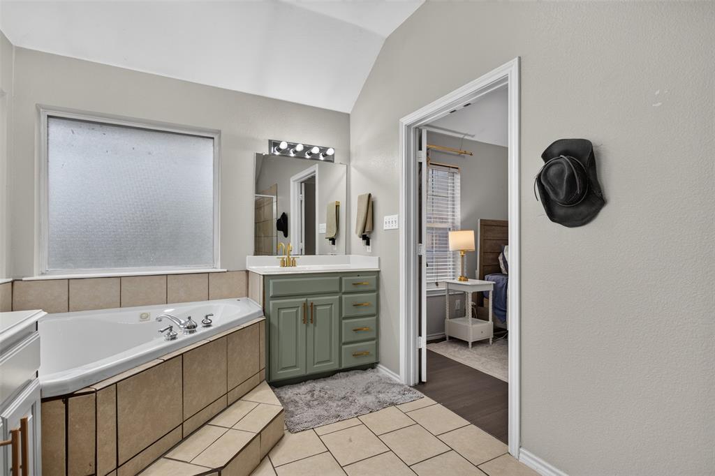 100 Radecke Road Krum, TX 76249 - Photo 26 of 29 Soaking tub with tiled surround, light-toned tile flooring, dual-basin vanity with a light green cabinet finish and gold-toned hardware, and a vaulted ceiling