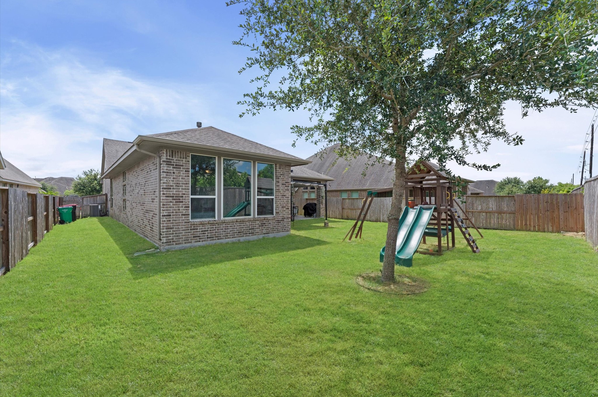 3510 Whitman Drive Rosharon, TX 77583 - Photo 18 of 25 This photo shows a spacious backyard with green grass, a tree, and a playset with slides. The brick house features large windows, and the area is enclosed by a wooden fence, providing a private outdoor space.