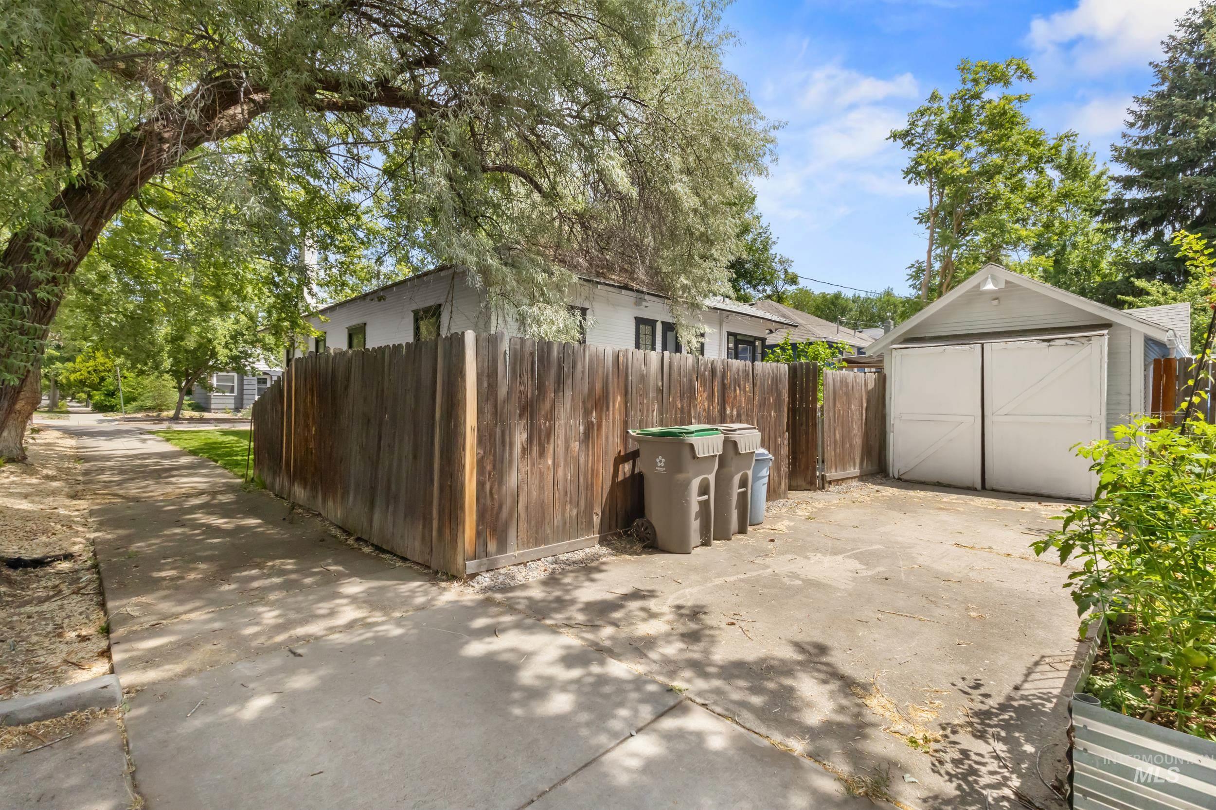 2119 West Brumback Street Boise, ID 83702 - Photo 27 of 30 Property exterior featuring a wooden privacy fence, a concrete driveway, a separate garage structure with double white doors, and mature trees providing natural canopy