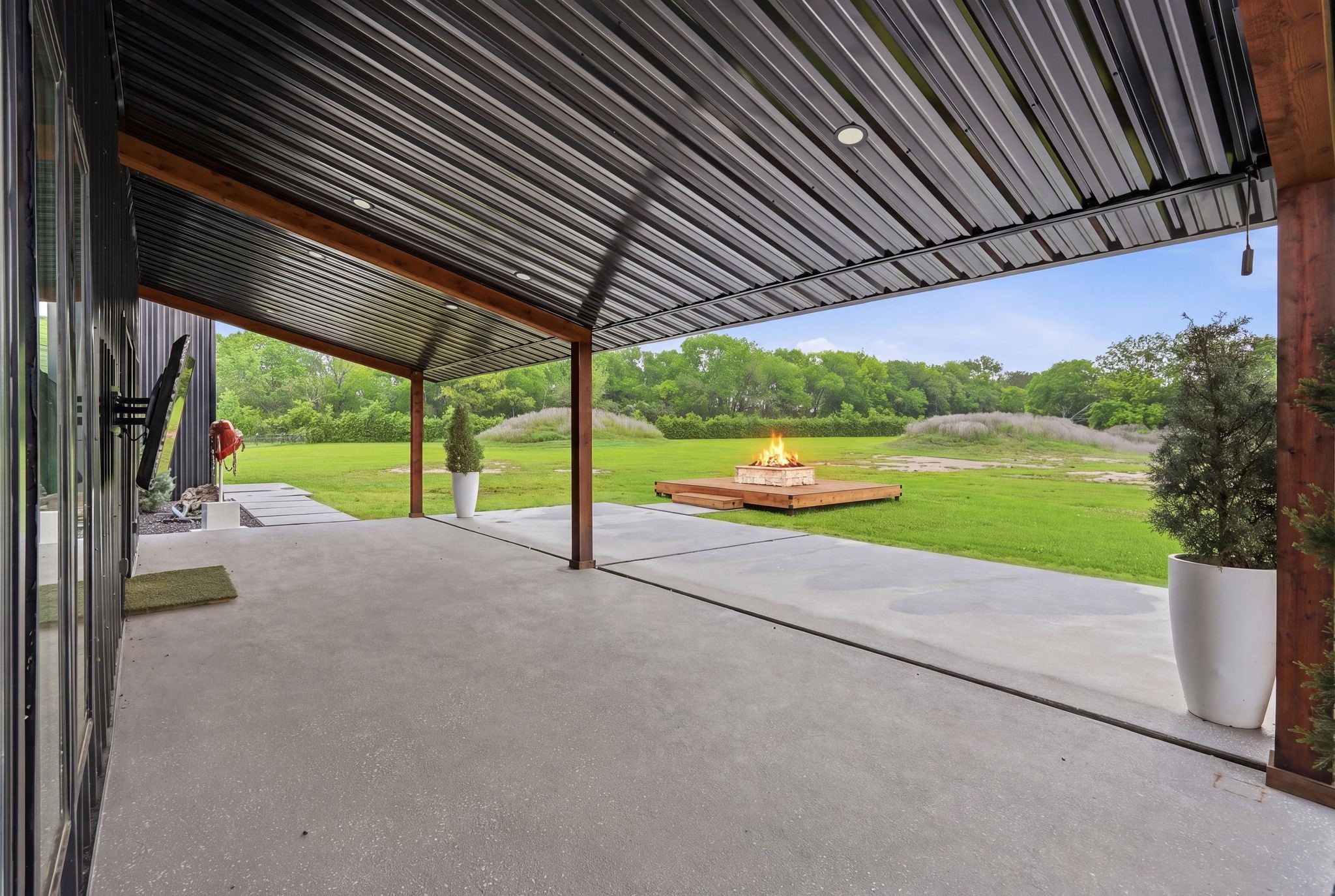 2743 Bobville Road Montgomery, TX 77316 - Photo 45 of 50 This photo showcases a spacious covered patio with a sleek metal roof, overlooking a lush green backyard. There's a modern fire pit centerpiece, perfect for outdoor gatherings, and large planters add a touch of greenery. Ideal for relaxing and enjoying the natural setting.