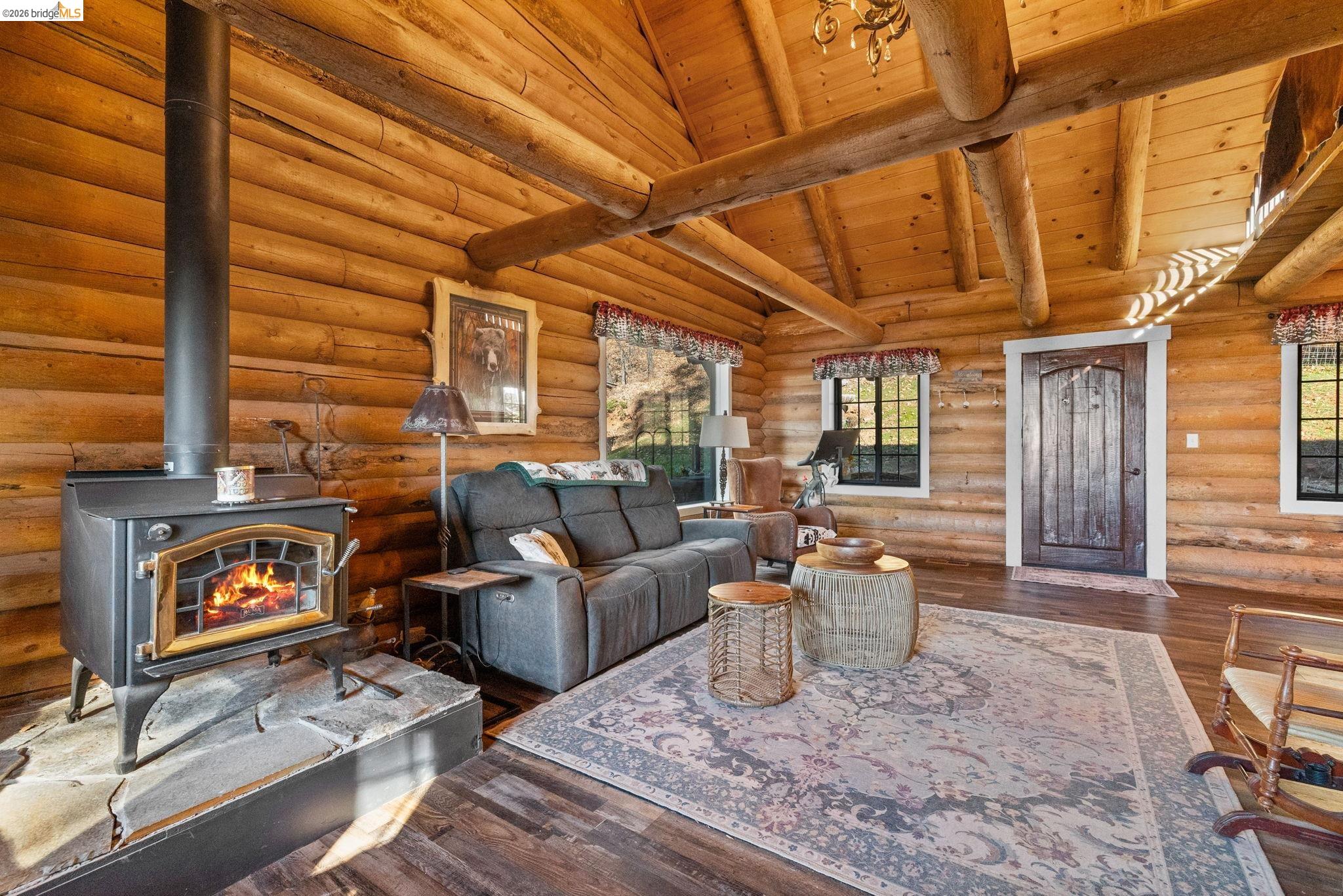 16144 Big Hill Road Sonora, CA 95370 - Photo 10 of 51 Vaulted ceiling featuring exposed log beams, wood-finish flooring, and a freestanding wood-burning stove with a stone hearth