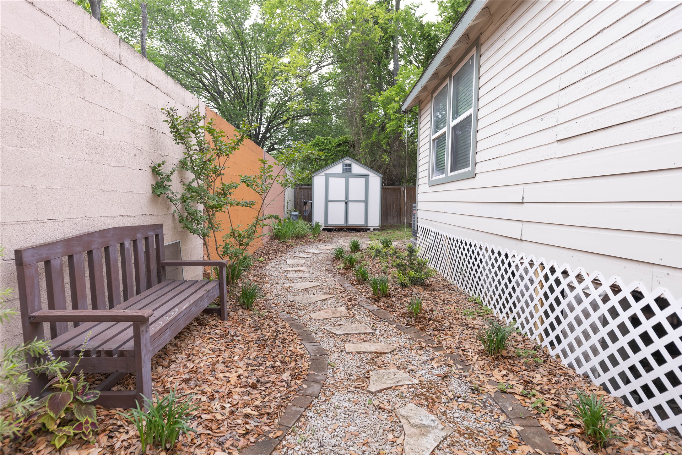 8007 Capitol Street Houston, TX 77012 - Photo 5 of 25 This photo showcases a narrow side yard featuring a stone pathway lined with mulch and small plants. A wooden bench sits against a concrete wall, and a white shed is visible at the end of the path, adjacent to a house with siding and lattice skirting.