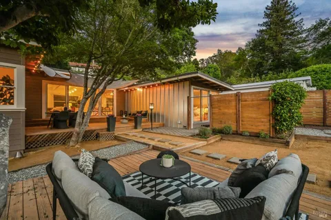 a view of a patio with couches table and chairs under an umbrella with large trees
