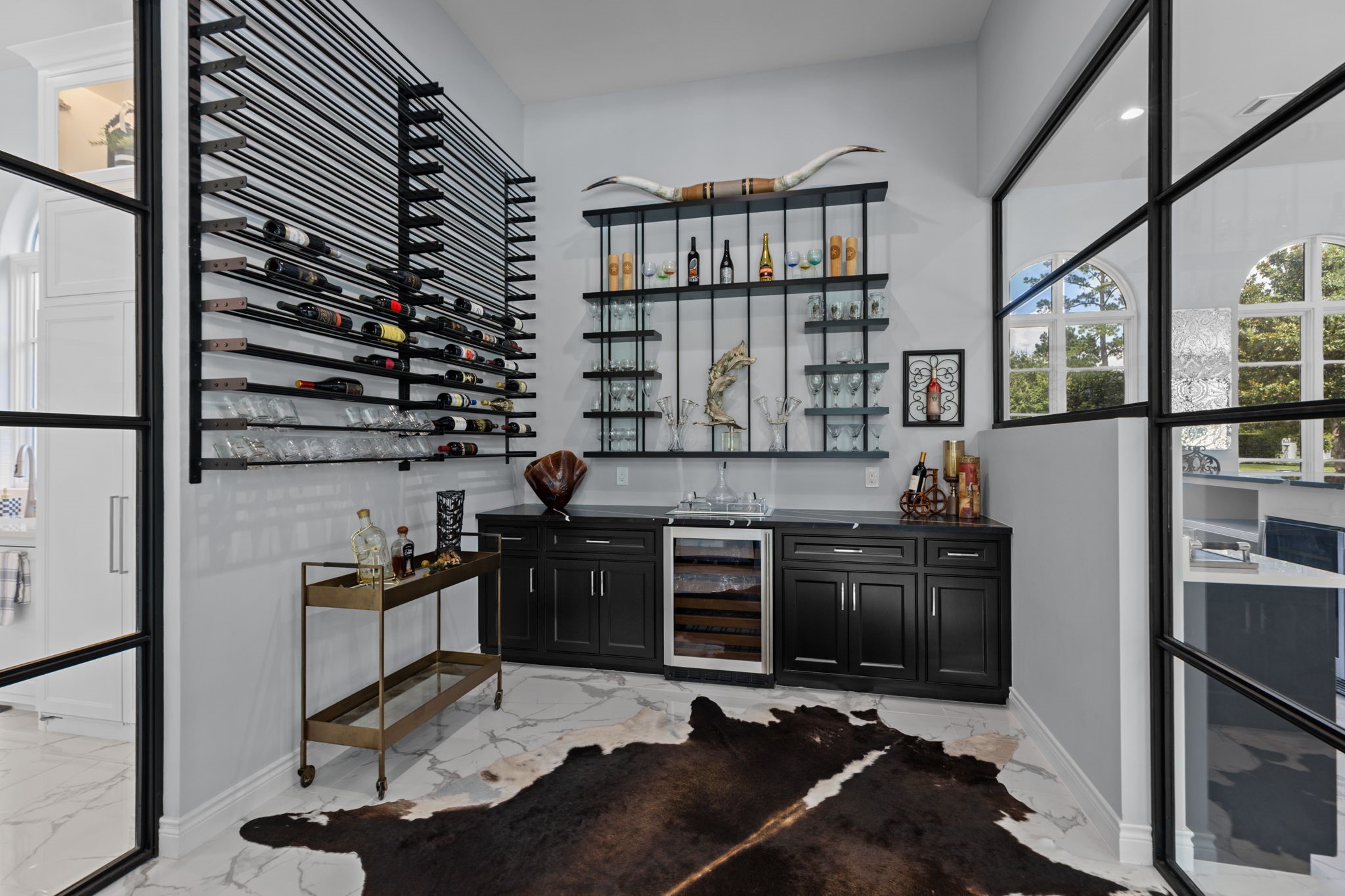 39 Maymont Way Spring, TX 77382 - Photo 11 of 50 This elegant home bar features sleek black cabinetry, a modern wine rack, and glass shelving. It includes a built-in wine cooler, a stylish bar cart, and a chic marble floor with a cowhide rug for added luxury.