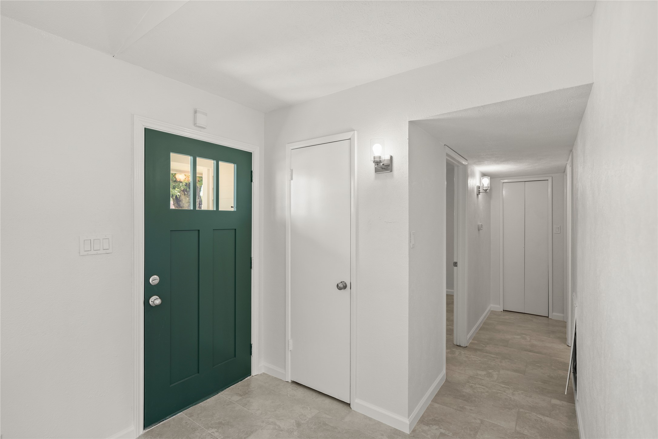6610 Blue Ridge Drive Richmond, TX 77469 - Photo 2 of 24 This photo showcases a bright entryway featuring a dark green door with windows, neutral tile flooring, and white walls. The space is well-lit with wall sconces and leads to a hallway with multiple bedrooms, offering a clean and inviting first impression.