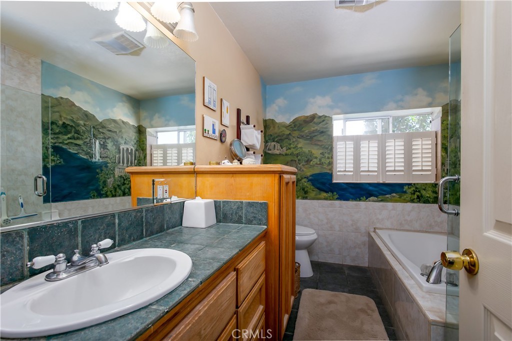 10960 Steinhoff Road Frazier Park, CA 93225 - Photo 32 of 46 The primary en-suite bathroom offers a functional retreat, complete with a long oak vanity with dual-sink capability, tile countertops, and a separate glass-enclosed shower. A highlight of the space is the deep jetted soaking tub, perfect for unwinding, set against a custom painted mountain-and-lake wall mural.