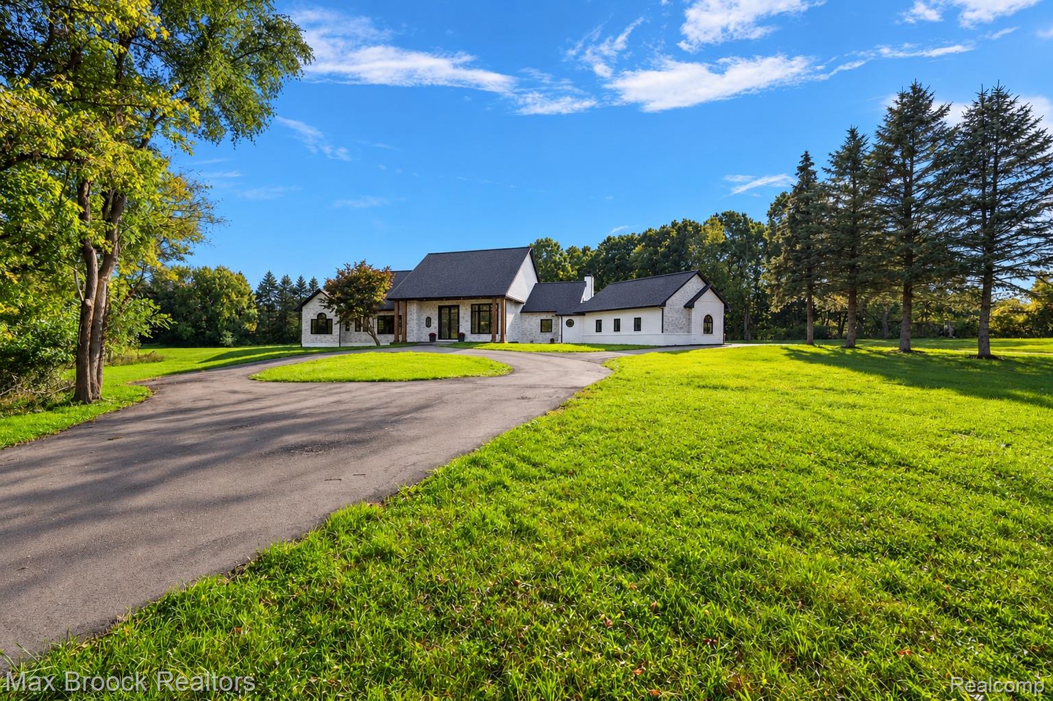 Spacious property featuring a circular driveway, expansive manicured lawn, mature trees, and a contemporary-style residence with a dark roof and white exterior