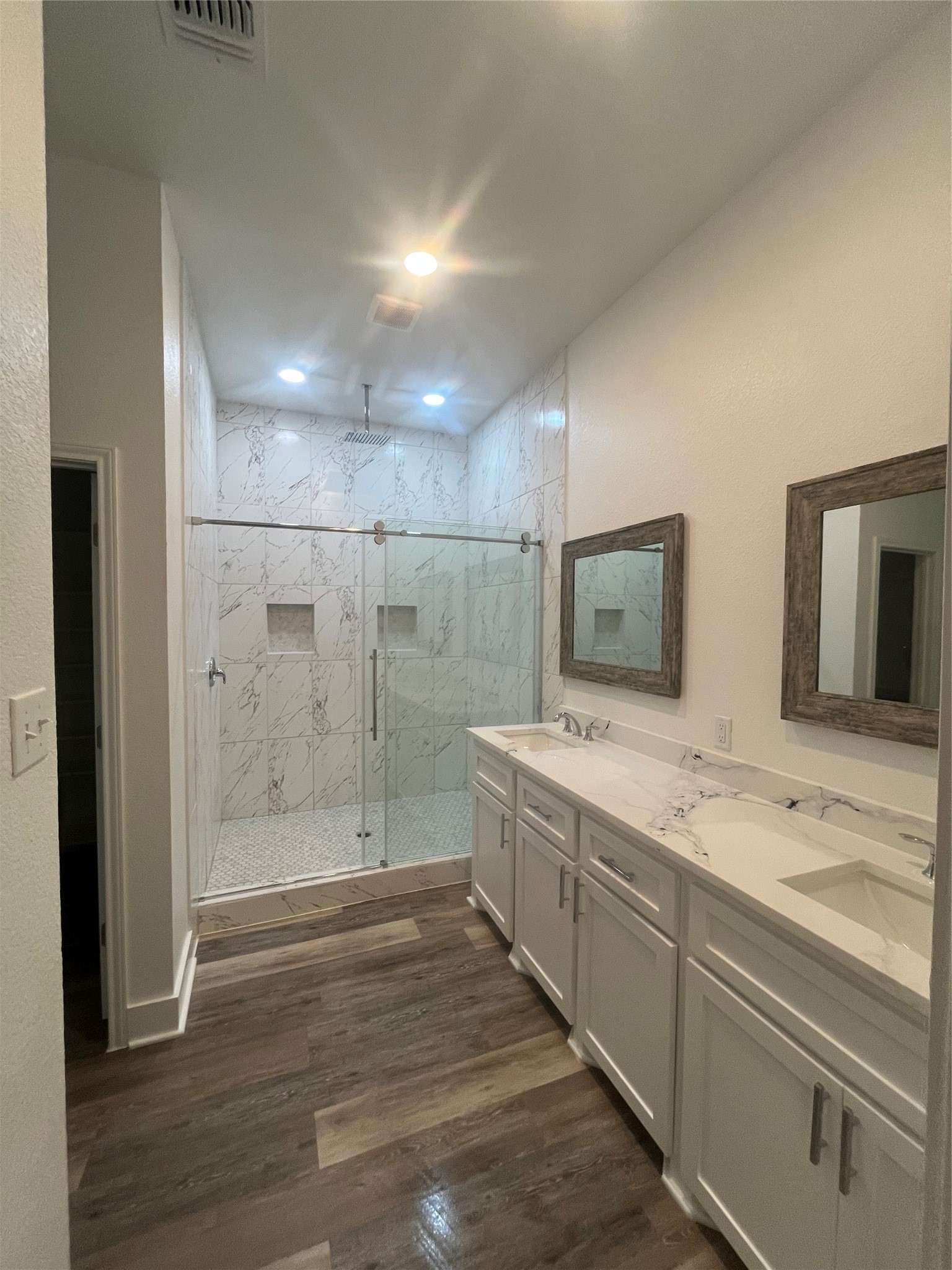 33416 FM 521 Road Angleton, TX 77515 - Photo 15 of 21 This bathroom features a spacious glass-enclosed shower with elegant tilework and dual shower niches. It includes a long, double-sink vanity with Quartz countertops and framed mirrors. The medium gray/wood water proof plank flooring adds warmth, and recessed lighting provides a bright, modern atmosphere.