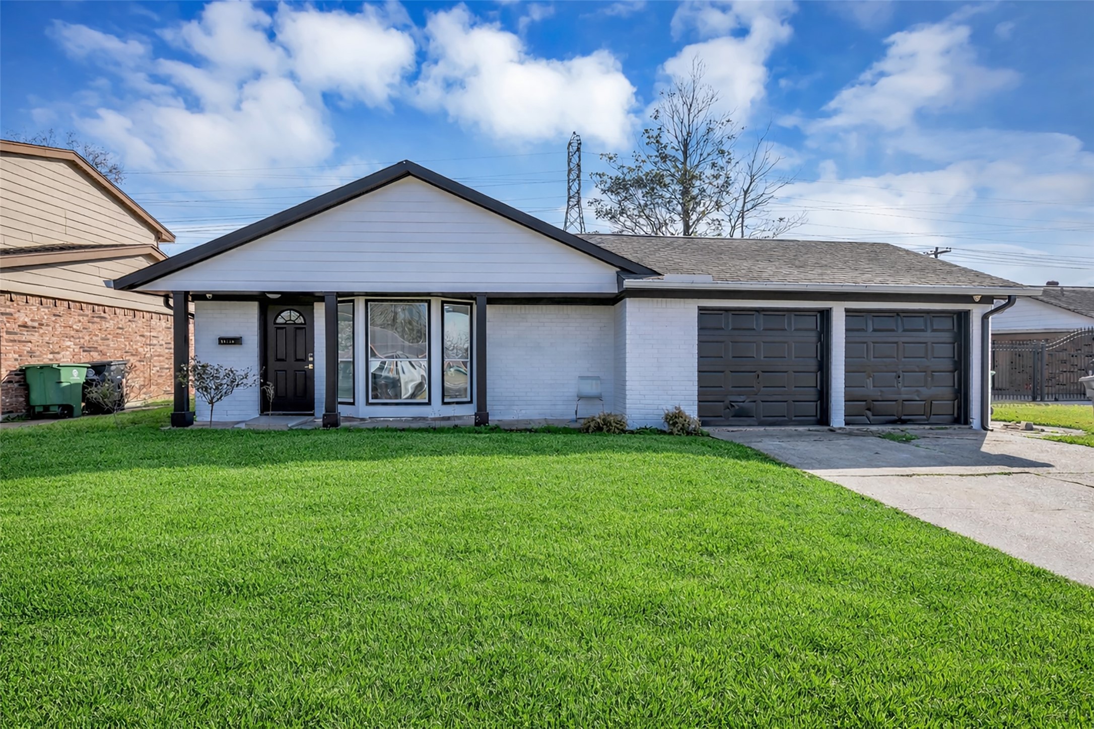 This is a single-story home with a clean, white exterior and black accents. It features an attached garage, a tidy front lawn, and a small porch with decorative columns. The windows are large, offering plenty of natural light.