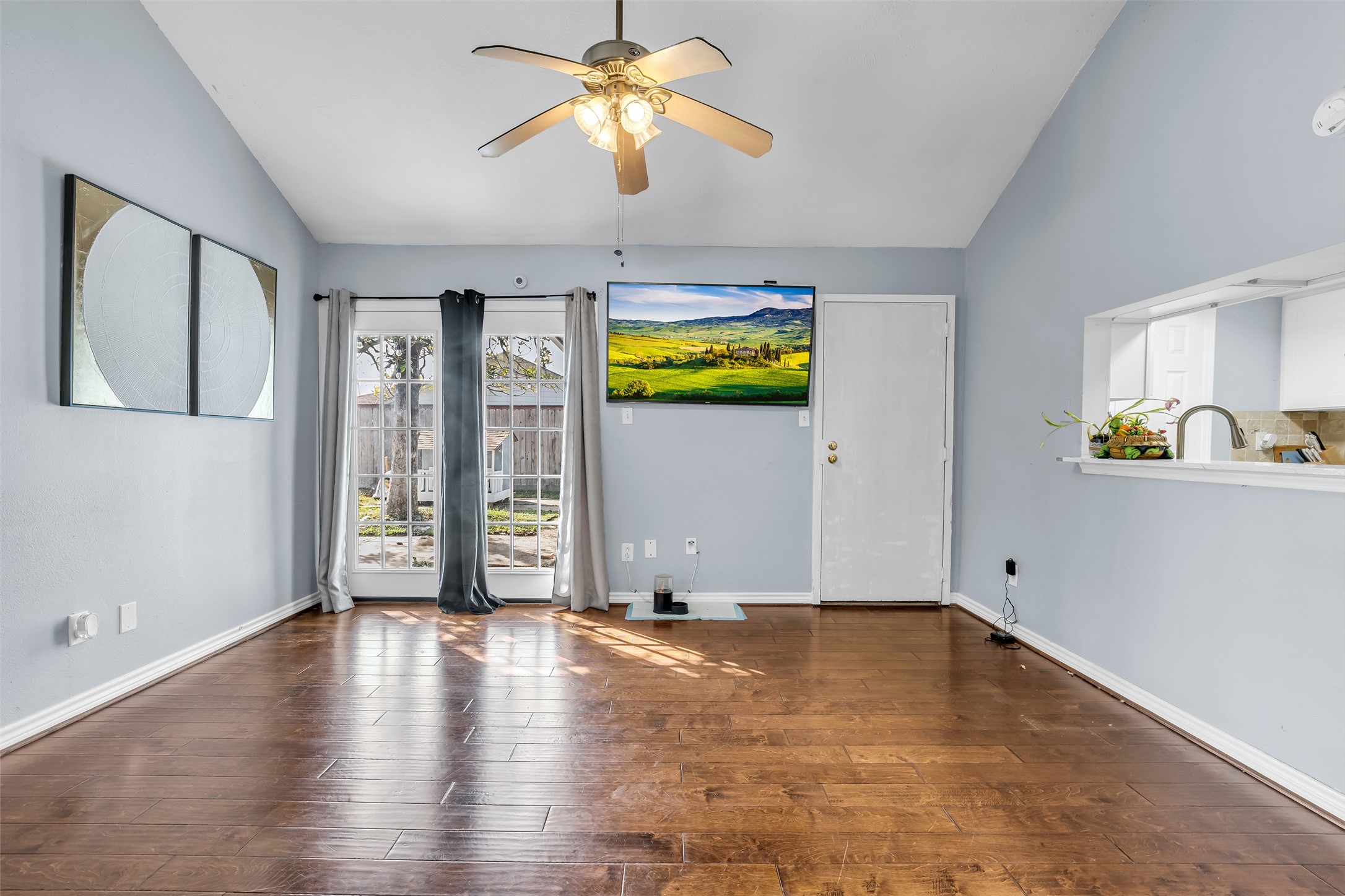 10730 Filey Lane Houston, TX 77013 - Photo 11 of 25 Bright living area with hardwood floors, featuring a ceiling fan and a large windowed door leading to the outdoors. A wall-mounted TV and open kitchen pass-through add modern touches. Neutral walls provide a versatile backdrop for any decor.