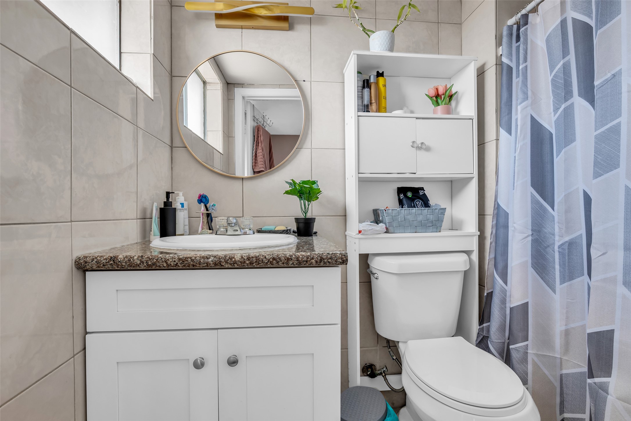 10730 Filey Lane Houston, TX 77013 - Photo 16 of 25 This bathroom features a clean, modern design with tiled walls, a round mirror above a vanity with granite countertop, and a white cabinet for storage. The space is brightened by a window.