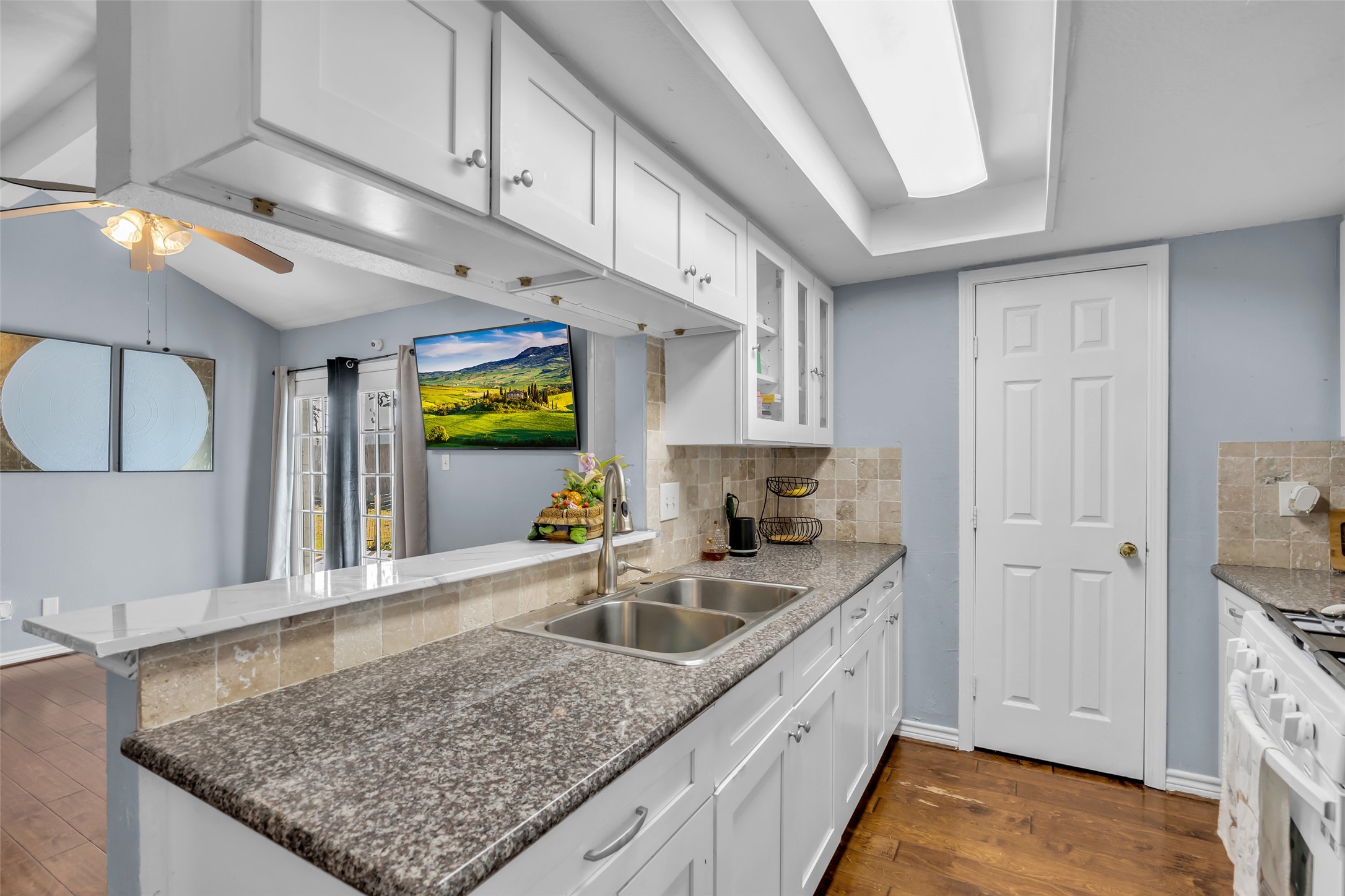 10730 Filey Lane Houston, TX 77013 - Photo 7 of 25 This photo showcases a modern kitchen with sleek granite countertops, white cabinetry, and a tiled backsplash. Natural light fills the space from a skylight, and there's an open view into the living area with hardwood floors and a ceiling fan.