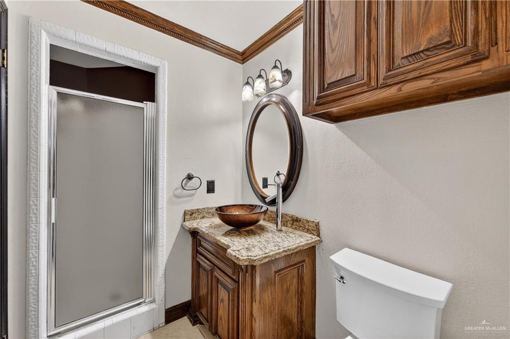 6004 North Cynthia Street McAllen, TX 78504 - Photo 12 of 25 Bathroom featuring a frameless shower enclosure with frosted glass, a wooden vanity with a granite-style countertop, a vessel sink, a wall-mounted oval mirror, and overhead wood cabinetry