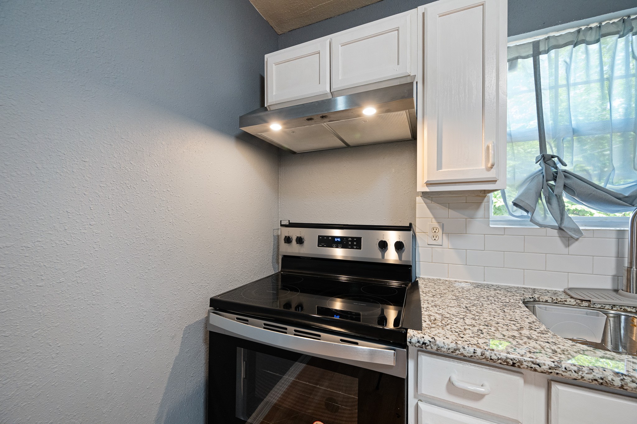 7815 Harding Street, Unit B Houston, TX 77012 - Photo 13 of 21 This modern kitchen boasts sleek granite countertops, a stylish electric range, and elegant white cabinetry, complemented by a sunlit window with charming curtains.