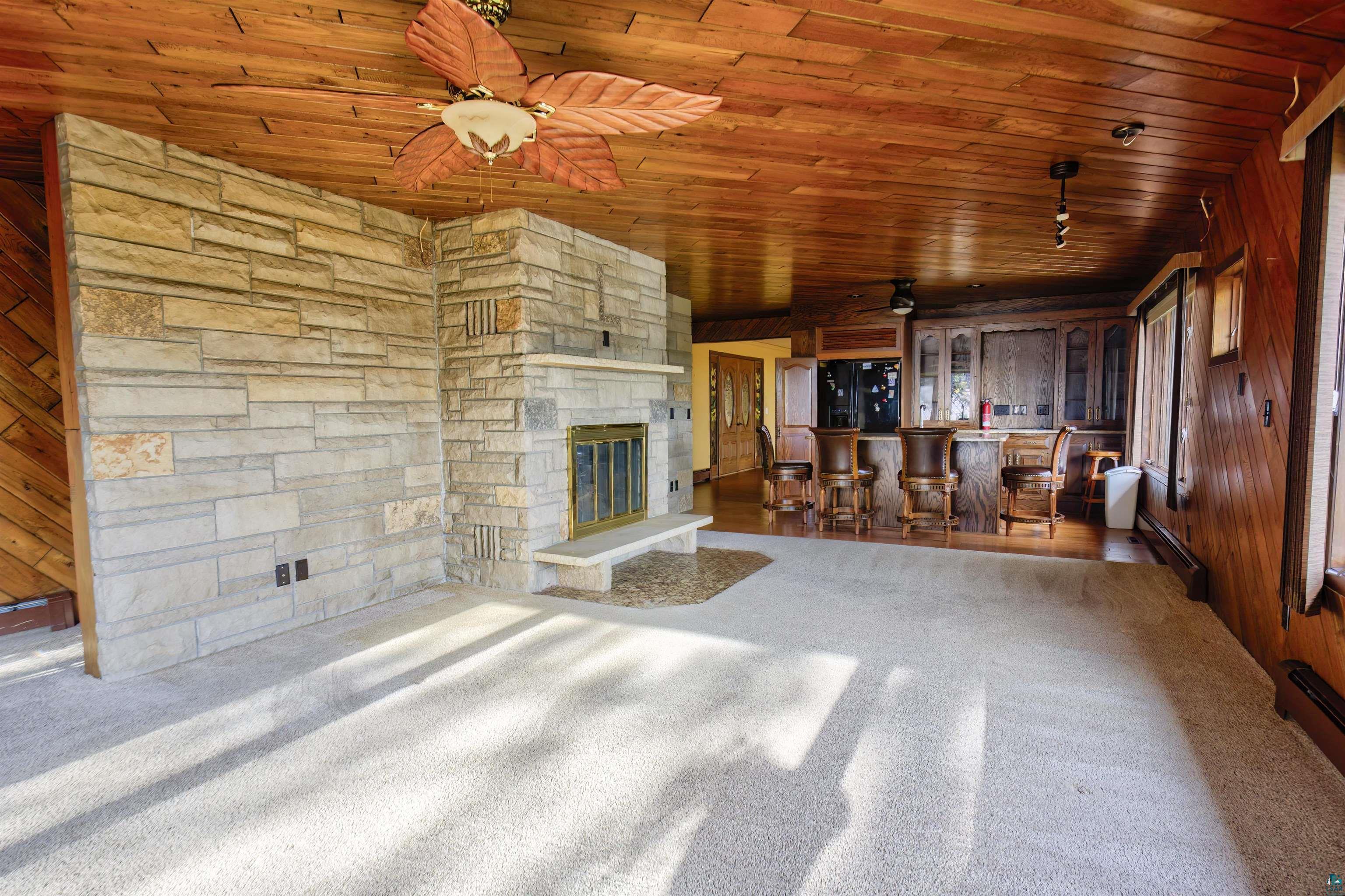 1933 West Shagawa Road Ely, MN 55731 - Photo 5 of 46 Unfurnished living room featuring a stone fireplace, ceiling fan, wooden ceiling, wooden walls, and indoor bar