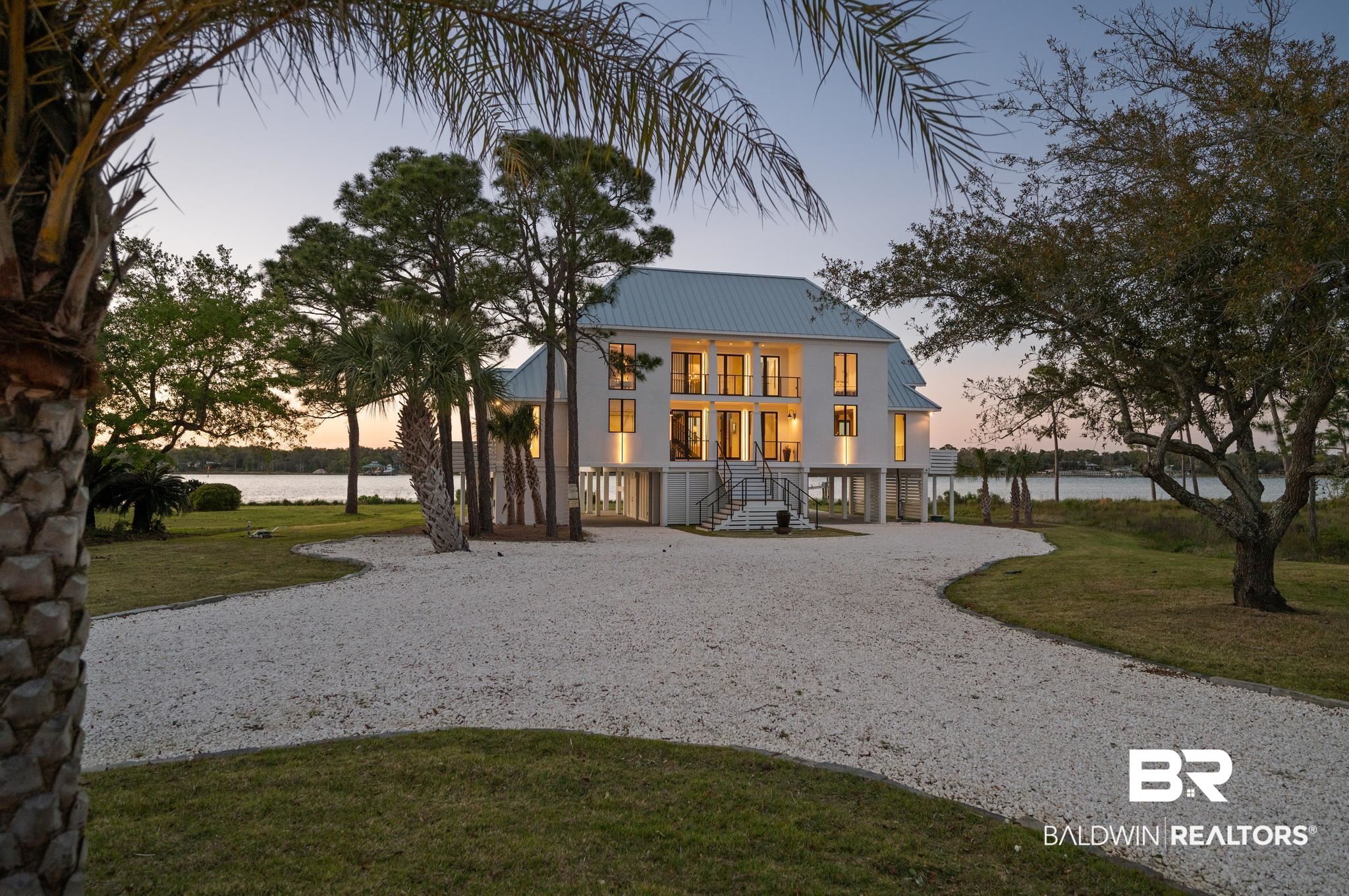 33349 River Road Orange Beach, AL 36561 - Photo 49 of 61 Waterfront residence featuring a light-colored exterior, multiple balconies, an expansive gravel driveway, mature trees, and a metal roof