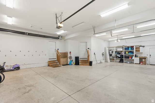 672 Perry Hollow Drive Mapleton, UT 84664 - Photo 38 of 106 Spacious garage featuring a light-toned concrete floor, multiple fluorescent light fixtures, two entry doors with wooden stairs, and built-in shelving units