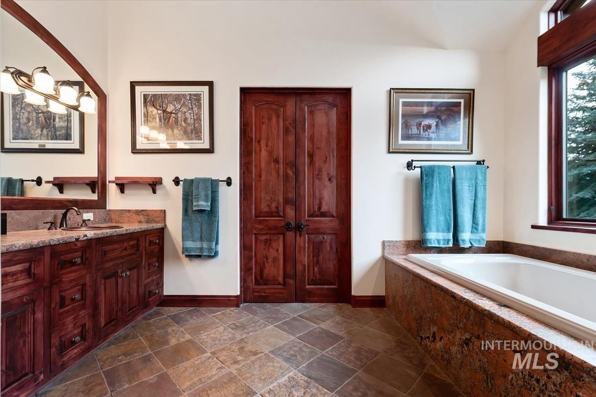 106 Hidden Hollow Road Hailey, ID 83333 - Photo 40 of 49 Rich wood vanity with integrated sink, stone countertop, spacious built-in tub with stone surround, and natural stone tile flooring
