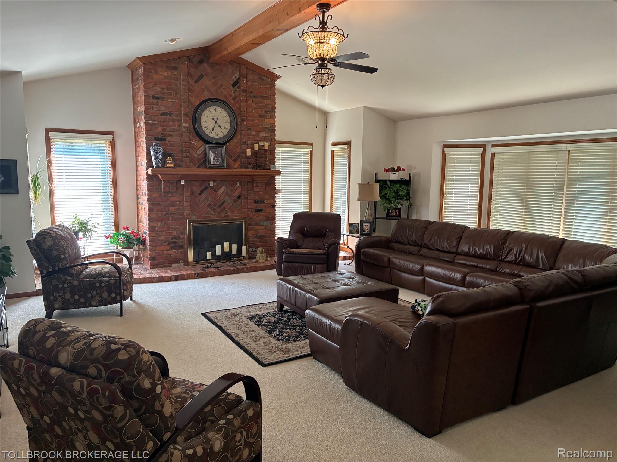 Vaulted ceiling with exposed wood beam, brick fireplace with wood mantel, multiple windows with blinds, and a built-in ceiling fan with integrated lighting