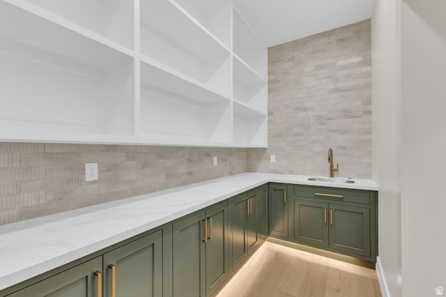 2828 Jojo Road Eagle Mountain, UT 84005 - Photo 13 of 63 Built-in cabinetry featuring white open shelving, a corner sink with a brass-finish faucet, dark green base cabinets with brass-finish hardware, and under-cabinet lighting, all complemented by light wood-finish flooring and a light-toned tiled backsplash