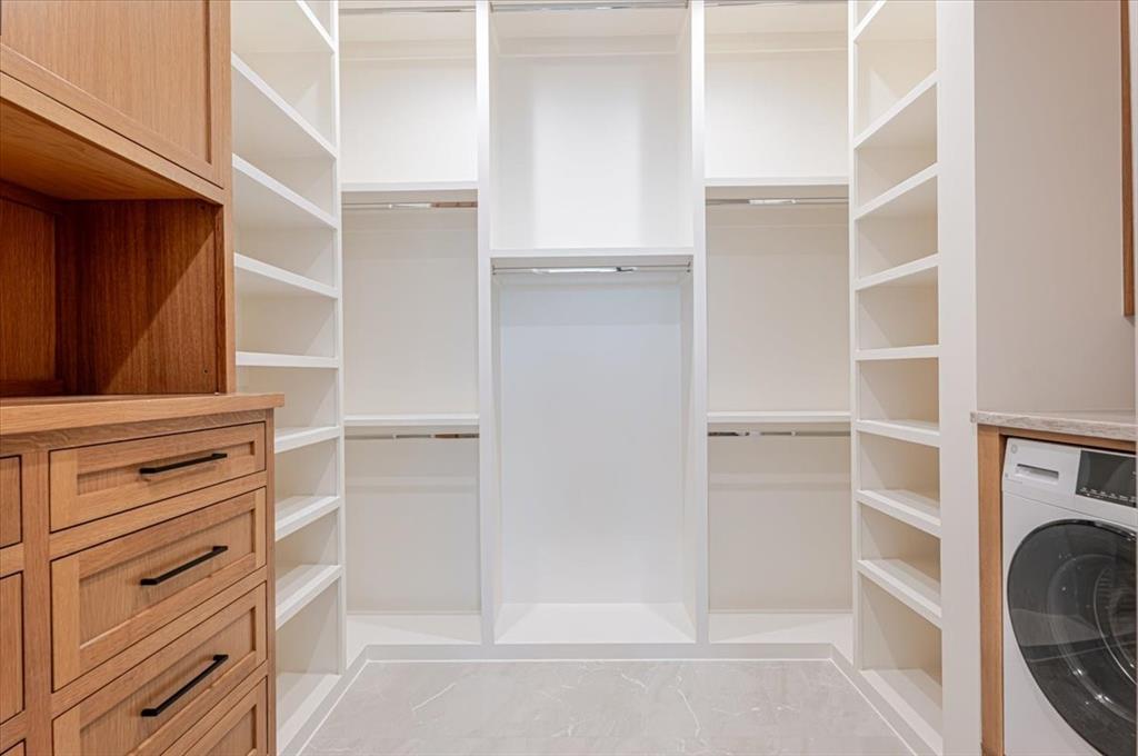 1014 Chaparrel Lane Graford, TX 76449 - Photo 15 of 40 Custom-Finished Primary Closet - Step into a beautifully appointed walk-in closet where thoughtful design meets everyday luxury. Custom two-tone cabinetry and open shelving provide generous, well-organized storage for wardrobes and accessories alike. A private in-suite laundry station with a sleek washer/dryer combo unit adds seamless convenience. The heated floor—extending from the bath into the closet—ensures comfort year-round. One of the home’s two attic access points is discreetly integrated here, preserving the clean architectural lines of the space.