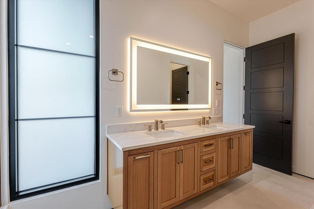 1014 Chaparrel Lane Graford, TX 76449 - Photo 22 of 40 Ensuite Bath with Smart Glass and Integrated Laundry - The ensuite guest bath blends convenience and comfort with a floating dual-sink vanity, backlit mirror, and heated towel bar. Full-height smart glass corner windows softly illuminates the space while preserving privacy. The walk-in closet is integrated into the bathroom area and features a private laundry station along with a second attic access—ideal for extended stays or added functionality.