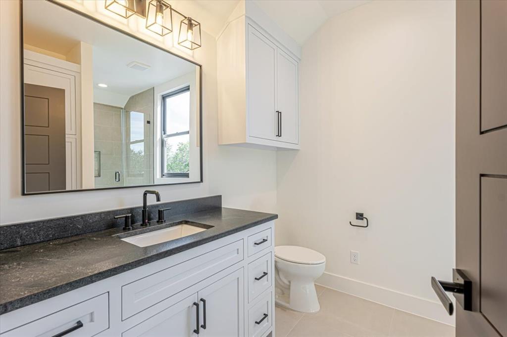 1014 Chaparrel Lane Graford, TX 76449 - Photo 29 of 40 The upstairs bathroom combines sleek modern design with practical storage. A spacious vanity with dark stone counters, generous drawers, and black fixtures sits beneath contemporary lighting and a large mirror. A walk-in tiled shower and private toilet area complete the space.