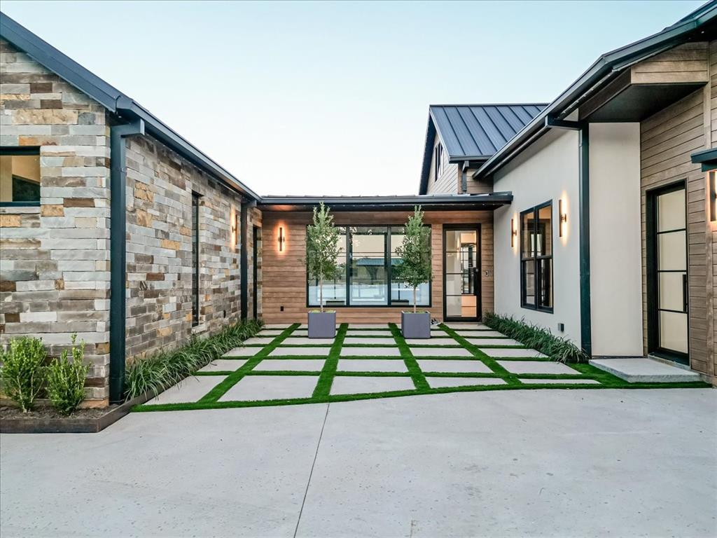 1014 Chaparrel Lane Graford, TX 76449 - Photo 5 of 40 Inviting Courtyard Entry - This thoughtfully designed courtyard blends clean architectural lines with warm natural materials. Large-format pavers set in artificial turf create a crisp, geometric pattern, flanked by horizontal wood-look steel siding, natural stone, and smooth stucco. Generous picture windows and a glass-paneled door frame the pool and lake view beyond, while integrated lighting, including under-eave aop-controlled Jellyfish lighting and symmetrical landscaping enhance the home’s modern elegance.
