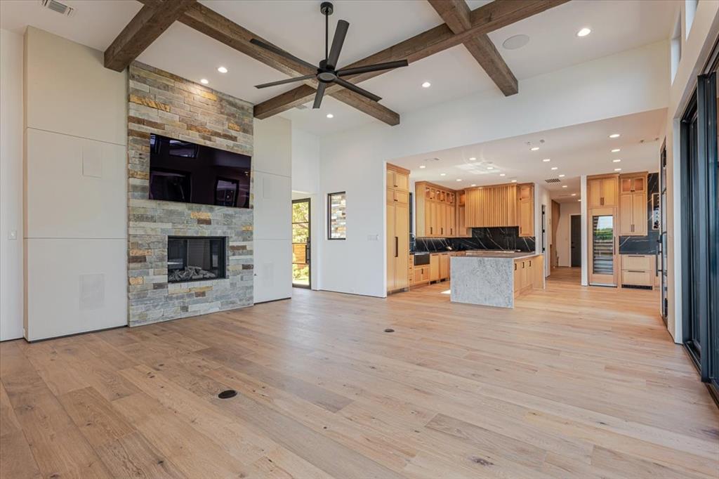 1014 Chaparrel Lane Graford, TX 76449 - Photo 6 of 40 Living Room - The heart of the home features wide-plank white oak flooring and striking wood beam accents that draw the eye upward. A floor-to-ceiling stone surround frames the gas log fireplace and mounted TV, creating a warm and refined focal point. Opposite to the fireplace, large sliding glass doors invite natural light and provide a stunning sightline to the lake, reinforcing the home’s seamless connection to its serene surroundings.