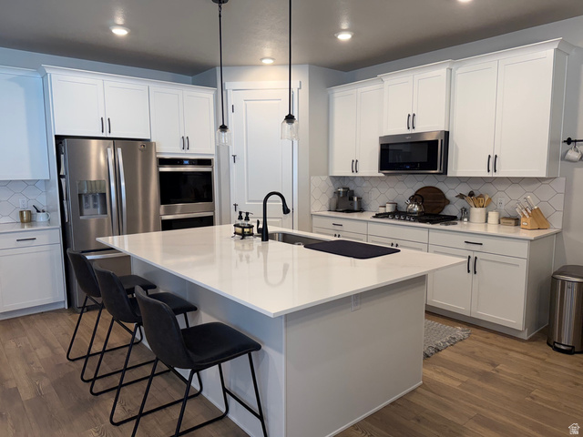 315 South Sherman Street Mapleton, UT 84664 - Photo 9 of 30 Bright kitchen featuring a substantial island with countertop seating, white shaker-style cabinetry, stainless steel appliances, a white geometric tile backsplash, and wood-finish flooring