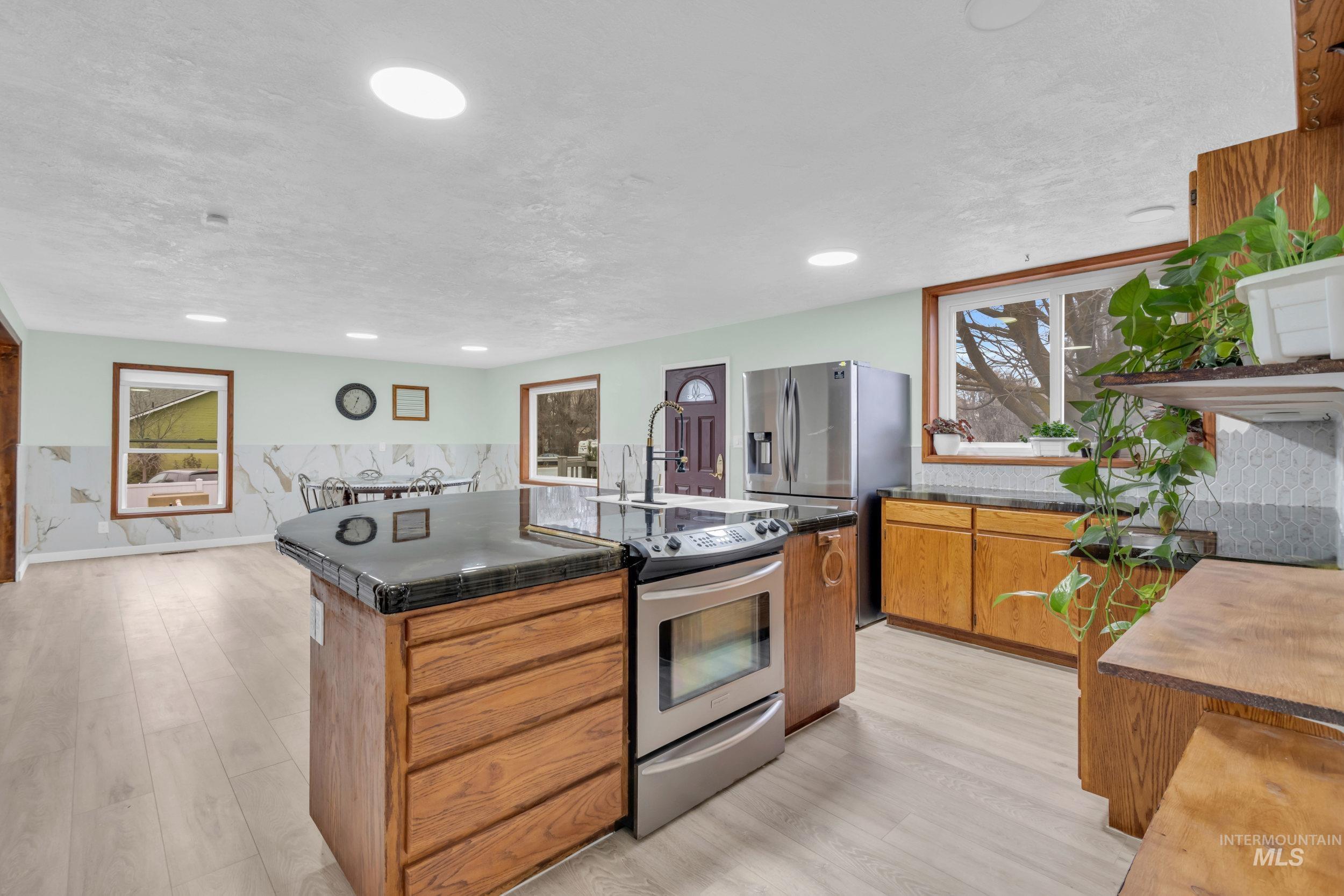 1158 South Powerline Road Nampa, ID 83686 - Photo 15 of 50 Kitchen with appliances with stainless steel finishes, brown cabinetry, a kitchen island with sink, dark countertops, and light wood finished floors