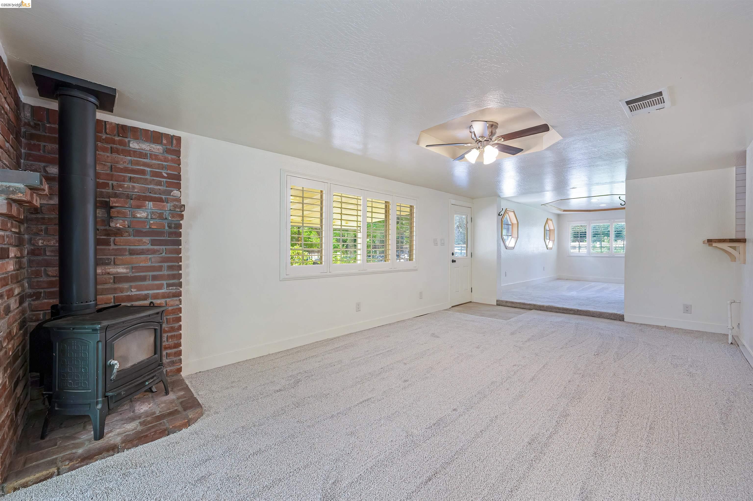 10474 Sheldon Road Elk Grove, CA 95624 - Photo 11 of 83 Spacious room featuring a brick wood-burning stove, light-colored carpeting, multiple windows with plantation shutters, a ceiling fan, and a built-in wall shelf