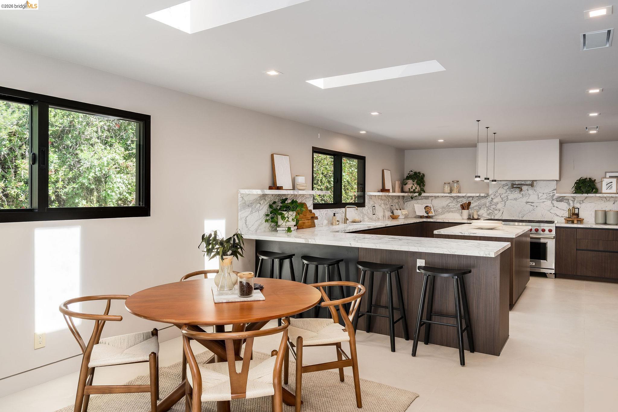 5645 Bacon Road Oakland, CA 94619 - Photo 37 of 94 Modern kitchen featuring a large central island with seating, dark wood-finish cabinetry, stone countertops with matching backsplash, stainless steel appliances, and skylights providing abundant overhead illumination
