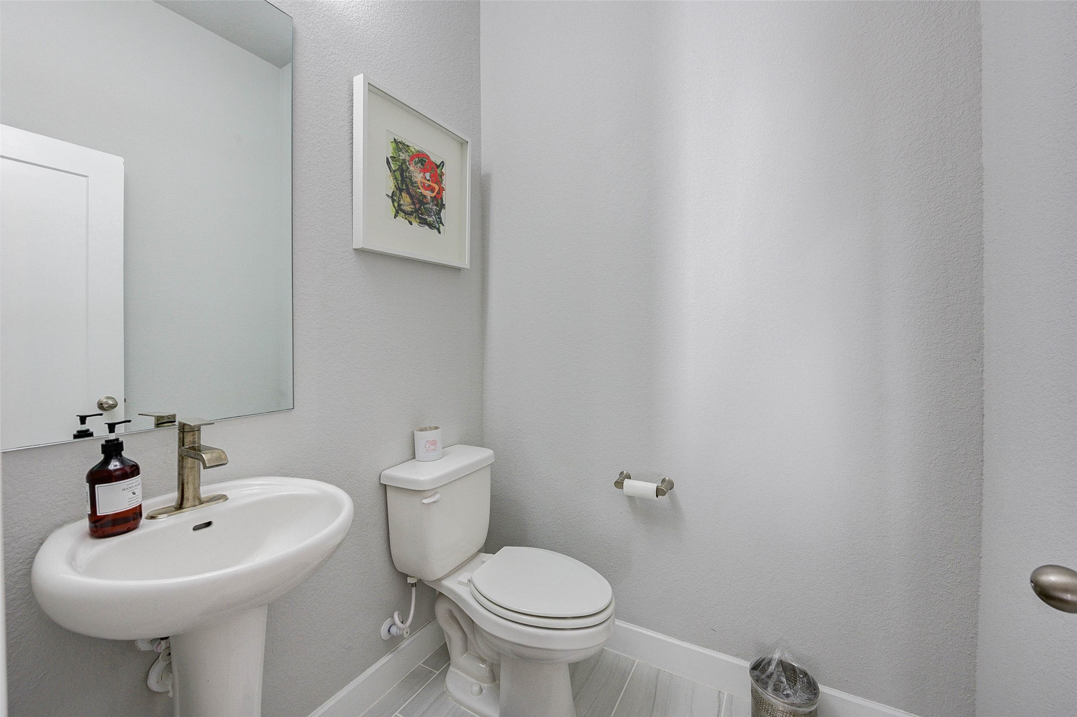 18732 Centro Main Street, Unit B Shenandoah, TX 77385 - Photo 16 of 42 This is a modern, compact half-bathroom featuring a pedestal sink, a toilet, and neutral-toned walls. It includes a large mirror, a small wall art piece, and has a clean, minimalistic design.