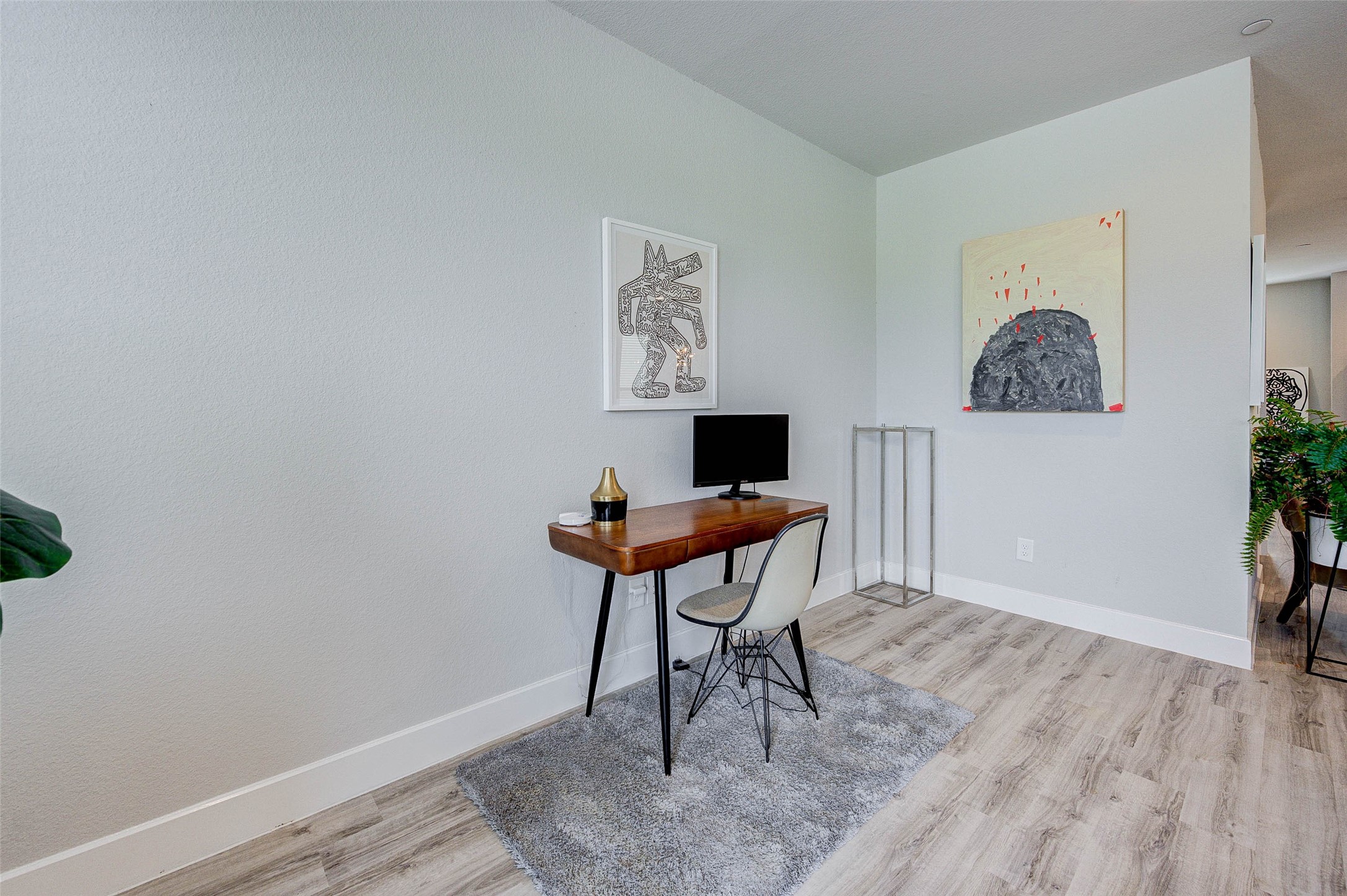 18732 Centro Main Street, Unit B Shenandoah, TX 77385 - Photo 19 of 42 This photo showcases a modern home office space with light wood flooring and neutral walls. It features a sleek wooden desk with a computer, a stylish chair, and contemporary artwork on the walls. The room is bright, minimalist, and inviting, perfect for a productive work environment.
