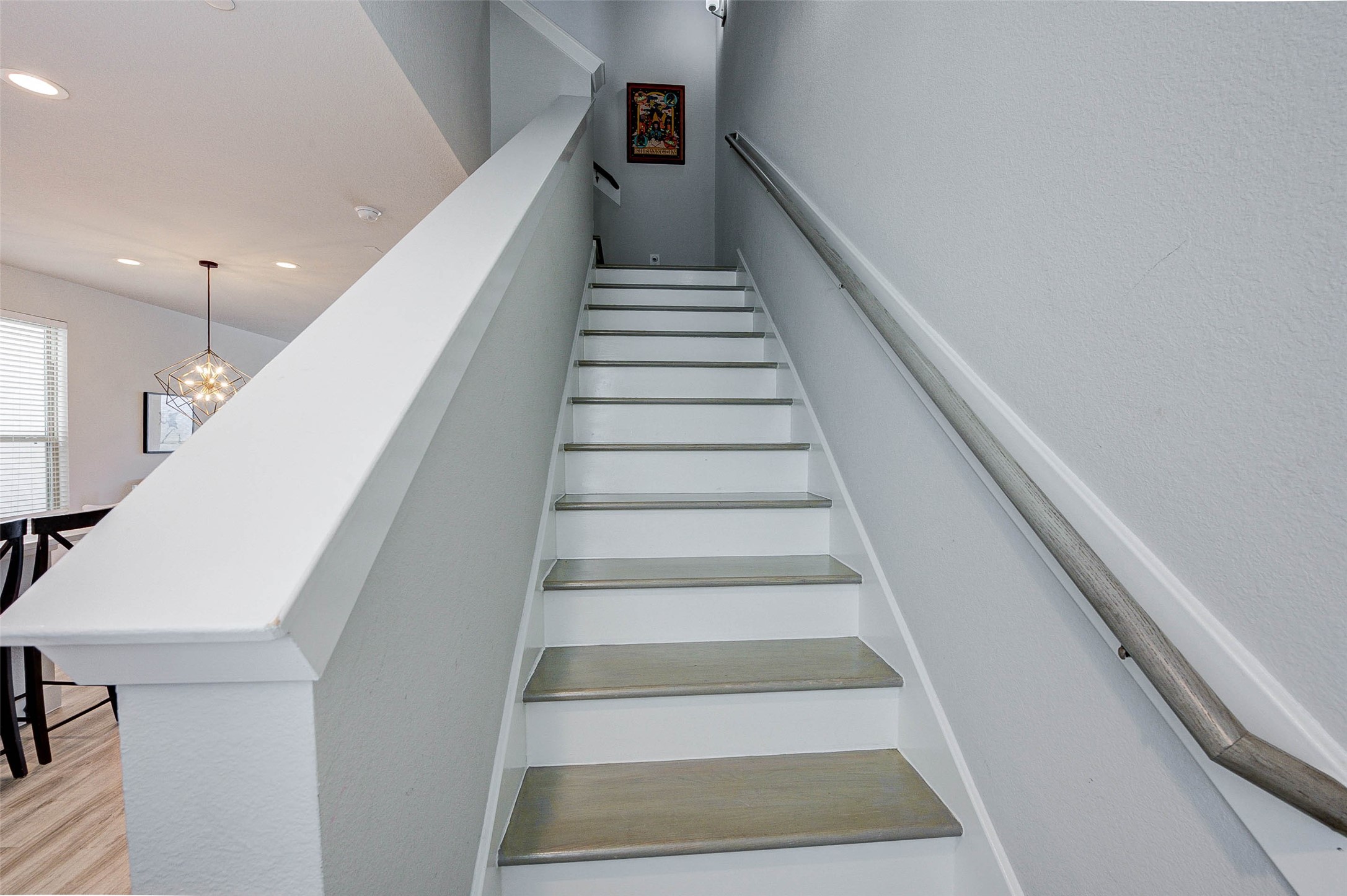 18732 Centro Main Street, Unit B Shenandoah, TX 77385 - Photo 23 of 42 This photo shows a modern staircase with light gray steps and a sleek handrail. The walls are painted a soft gray, and there's a glimpse of a dining area with contemporary lighting. A small piece of artwork hangs at the top of the stairs.