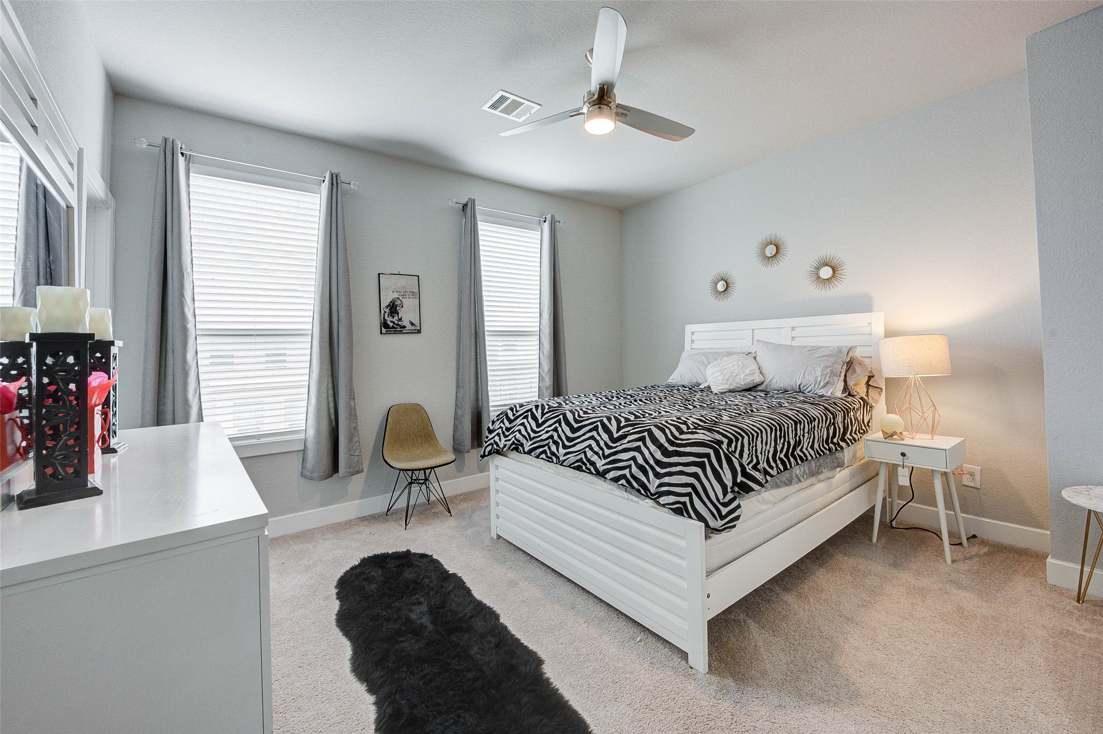18732 Centro Main Street, Unit B Shenandoah, TX 77385 - Photo 24 of 42 This bedroom features a cozy, modern design with soft carpet flooring and neutral walls. It includes a stylish bed with a zebra-patterned duvet, a ceiling fan, and ample natural light from two large windows with gray curtains. A chic dresser and nightstand, complemented by decorative wall accents, complete the room.