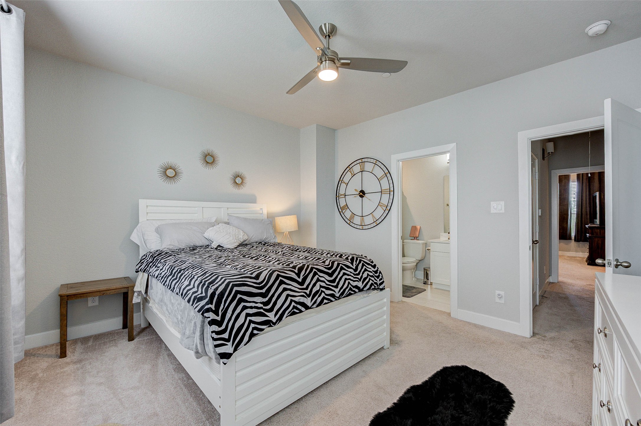 18732 Centro Main Street, Unit B Shenandoah, TX 77385 - Photo 25 of 42 This is a cozy and bright bedroom with a modern ceiling fan, neutral walls, and carpeted flooring. It features a large wall clock, stylish decor, and an en-suite bathroom, providing both functionality and style.