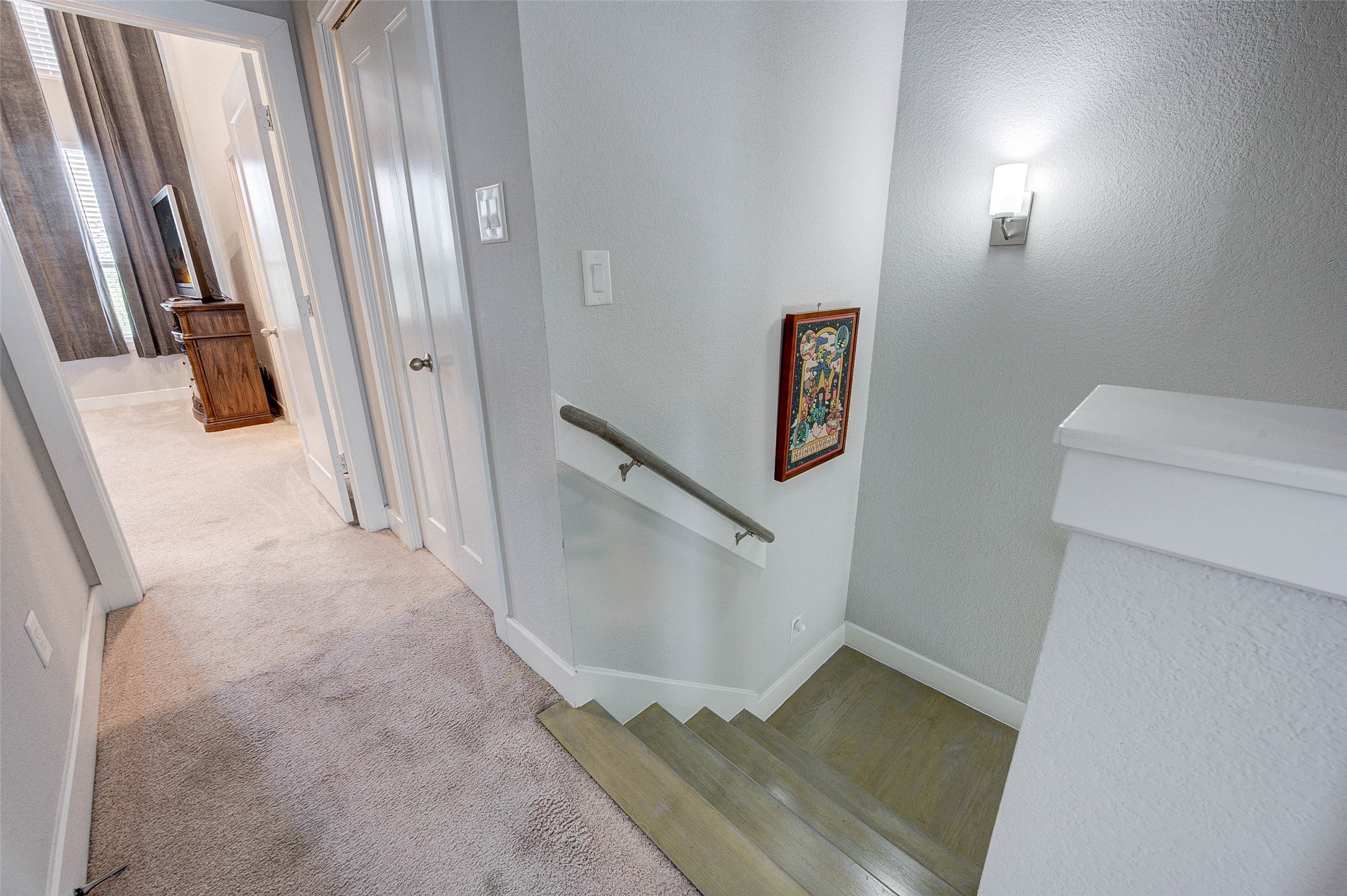 18732 Centro Main Street, Unit B Shenandoah, TX 77385 - Photo 27 of 42 This photo shows a bright, carpeted hallway leading to a bedroom with large windows and a TV. It features a well-lit stairway with a wooden handrail and a decorative wall piece.