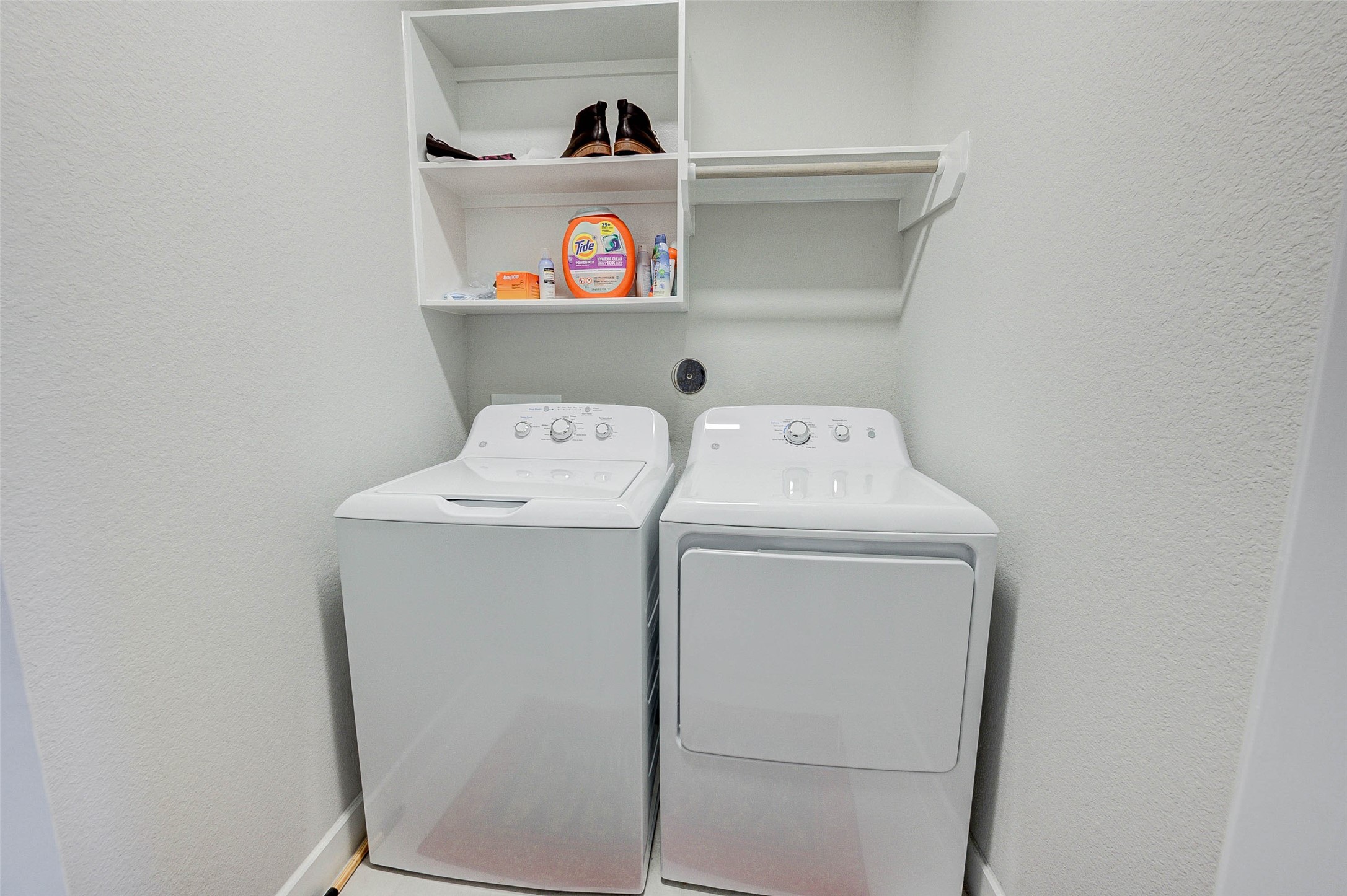 18732 Centro Main Street, Unit B Shenandoah, TX 77385 - Photo 28 of 42 This photo shows a compact laundry area with a washer and dryer side by side. Above the appliances, there's shelving for storage, ideal for keeping detergents and laundry essentials organized. The space is clean and efficiently designed.