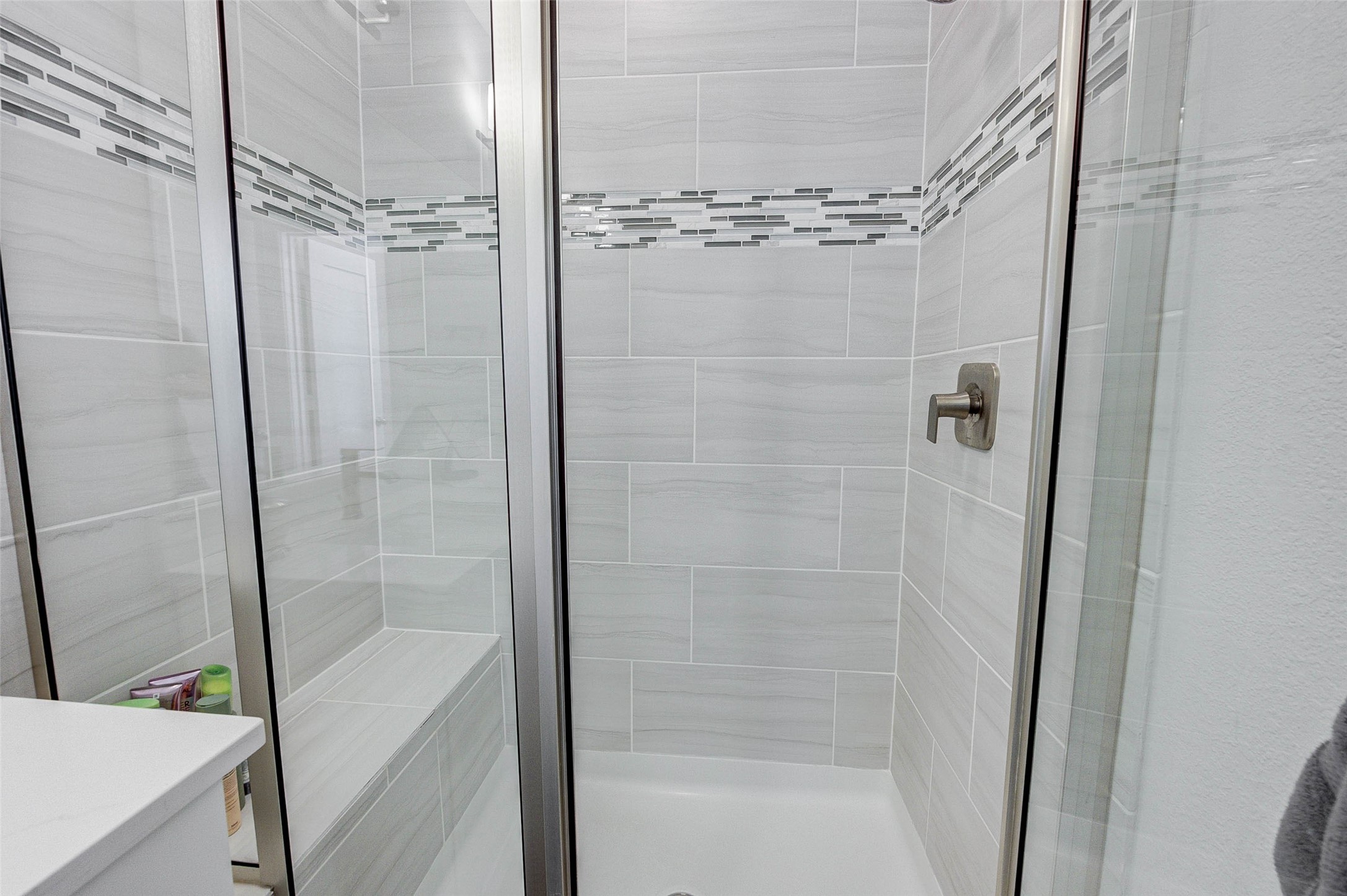 18732 Centro Main Street, Unit B Shenandoah, TX 77385 - Photo 33 of 42 This photo shows a modern shower with sleek glass doors and a built-in bench. The walls are lined with light gray tiles accented by a decorative band of smaller, darker tiles, creating a stylish and contemporary look.