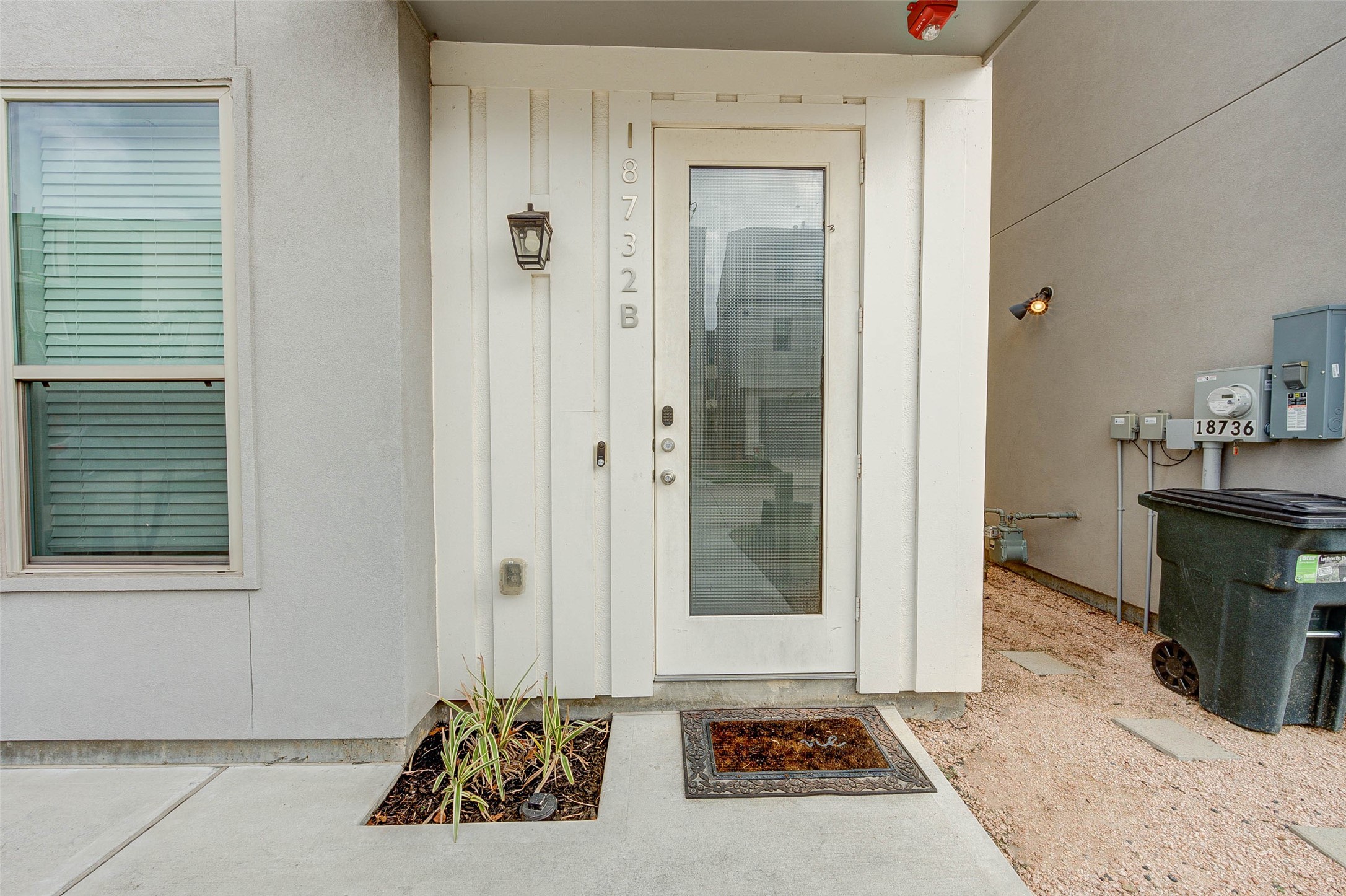 18732 Centro Main Street, Unit B Shenandoah, TX 77385 - Photo 38 of 42 This photo shows a modern entryway with a glass-panel door and a simple exterior light fixture. The area features a small planted bed and a concrete pathway, providing a clean and welcoming entrance.
