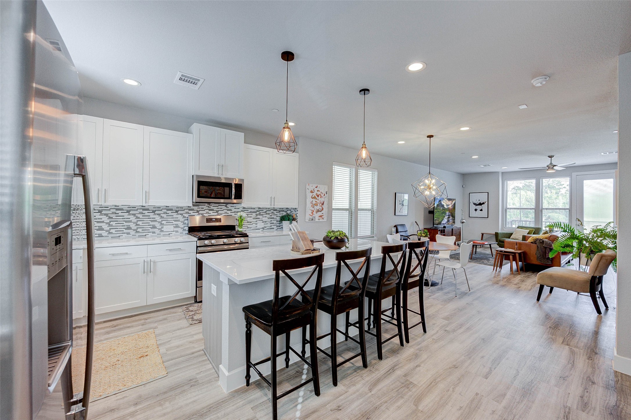 18732 Centro Main Street, Unit B Shenandoah, TX 77385 - Photo 8 of 42 Modern open-concept kitchen and living area with sleek white cabinetry, stainless steel appliances, and stylish pendant lighting. The space features a functional island with seating, light wood flooring, and ample natural light from large windows, creating a welcoming atmosphere.