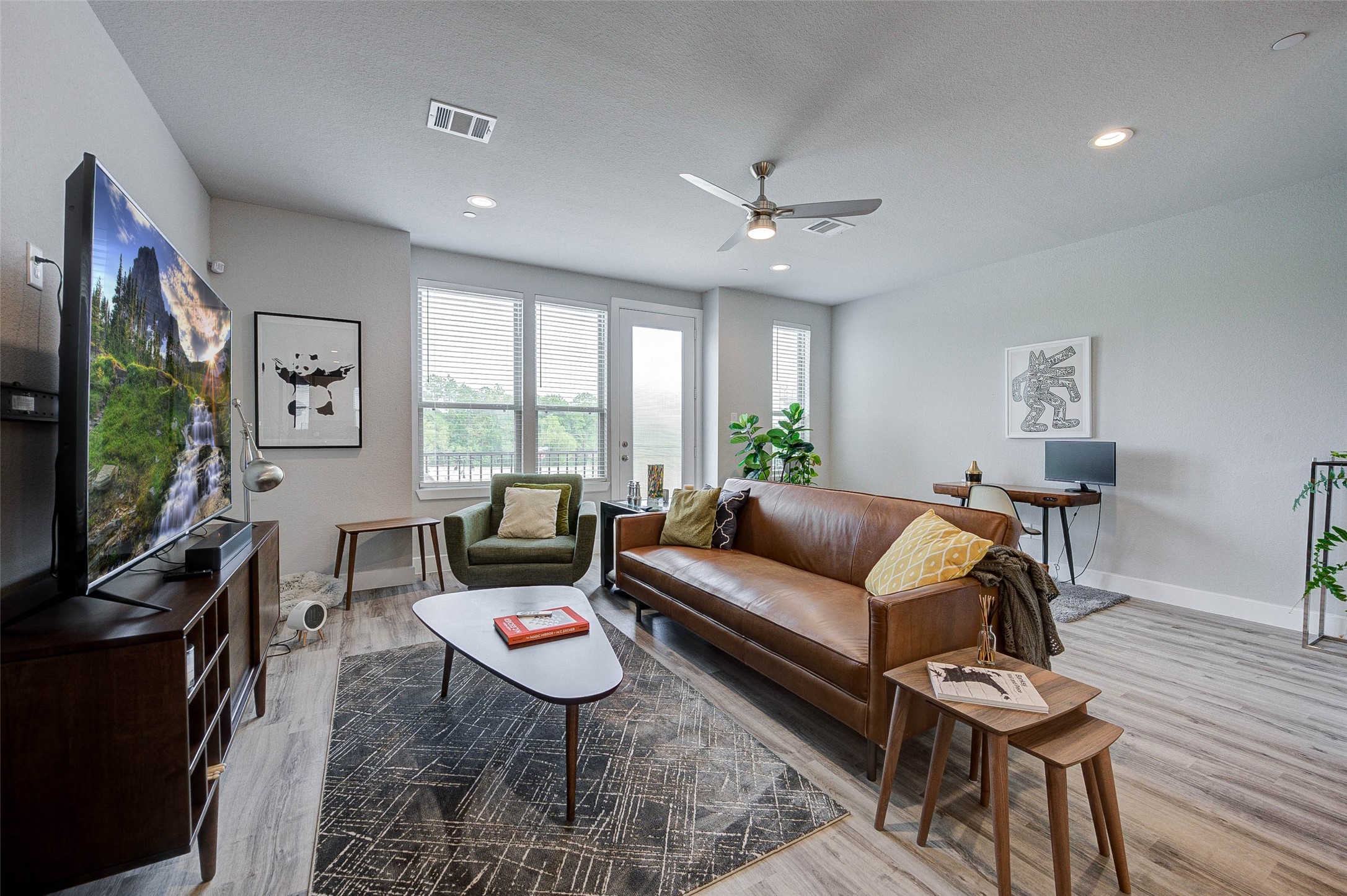 18732 Centro Main Street, Unit B Shenandoah, TX 77385 - Photo 10 of 42 Bright and modern living room with large windows, featuring a stylish leather sofa, sleek TV setup, and a small work desk. The space is accented with plants and contemporary artwork, offering a cozy yet sophisticated atmosphere.
