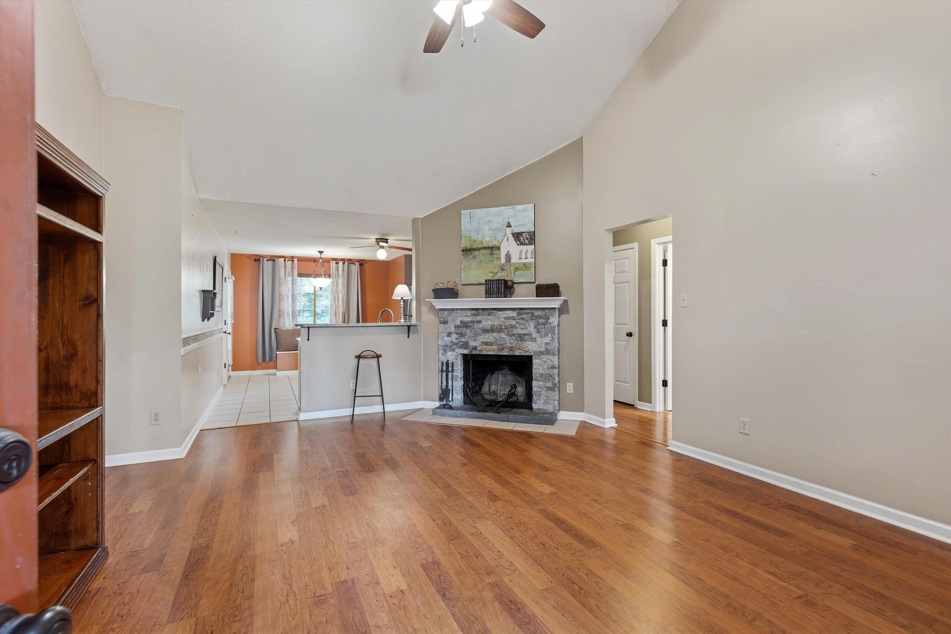 Living area featuring wood-finish flooring, a stone-faced fireplace with a mantel, and a vaulted ceiling with a ceiling fan