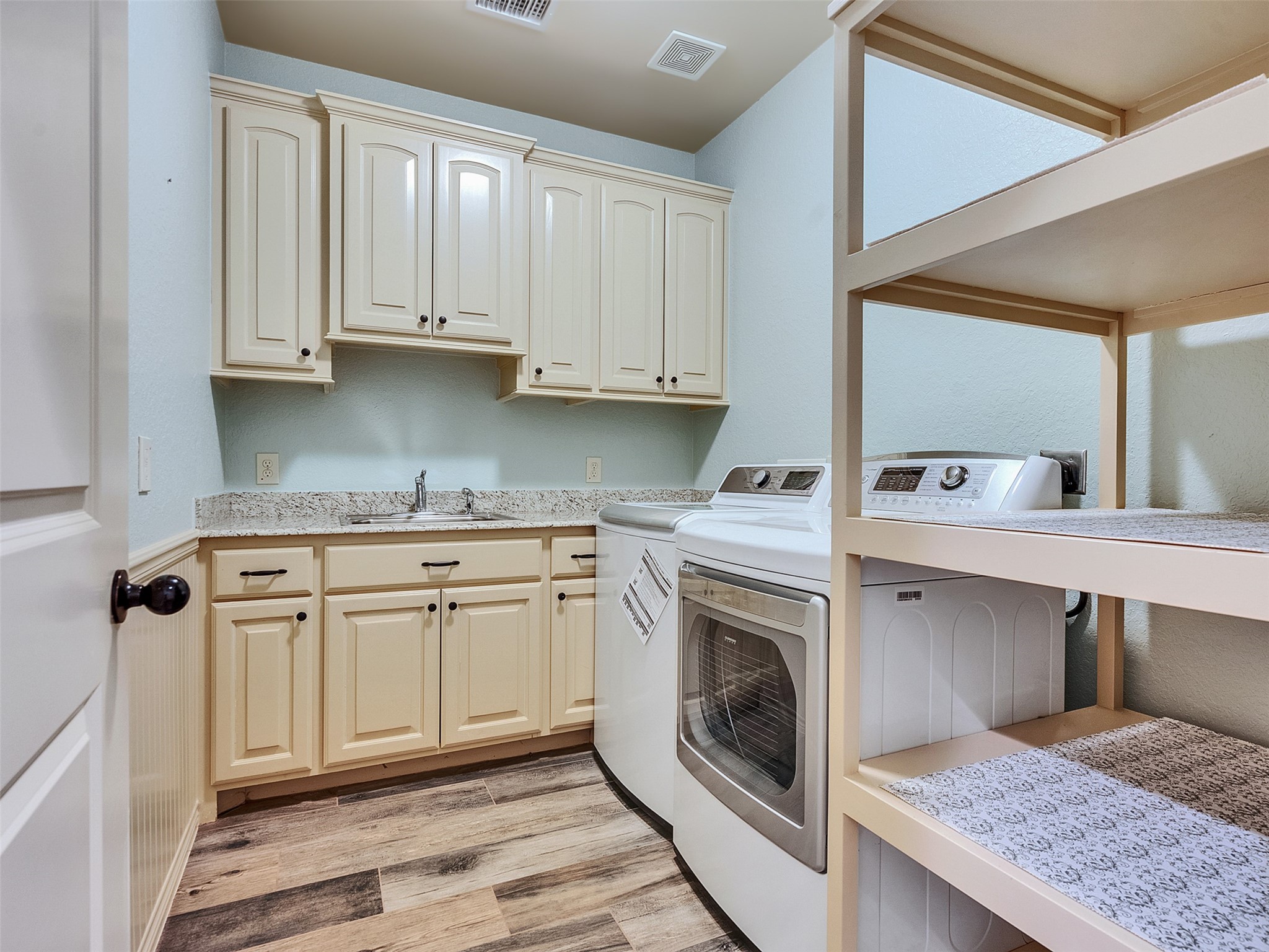 18005 Cindy Lane Conroe, TX 77302 - Photo 37 of 49 This photo showcases a well-organized laundry room featuring a washer and dryer, ample cabinetry for storage, a sink with a granite countertop, and open shelving. The space has a light color palette and wood-look flooring.